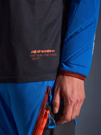 Primo piano della maglia Alpinestars Maxdura Dual in grigio scuro e blu reale, con logo arancione e scritta 'One Goal One Vision' sul busto in rete, manica imbottita con pannelli geometrici rinforzati, dettaglio tecnico per guida adventure
