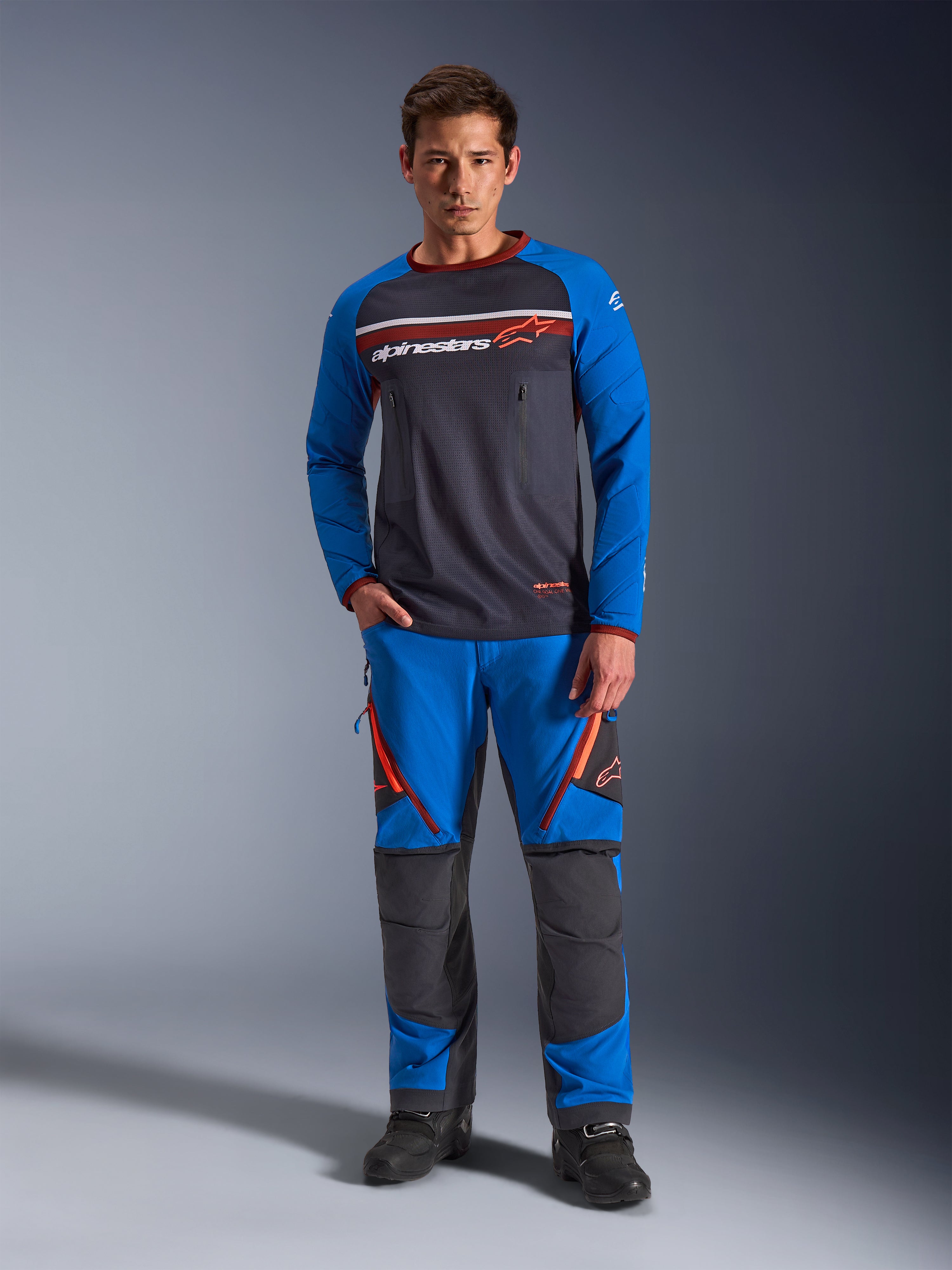 Alpinestars Maxdura Dual Jersey, maglia da moto, grigio scuro e blu reale, torso in rete con maniche imbottite e tasche sul petto con cerniera, logo Alpinestars bianco e arancione, indossata con pantaloni adventure abbinati e stivali neri