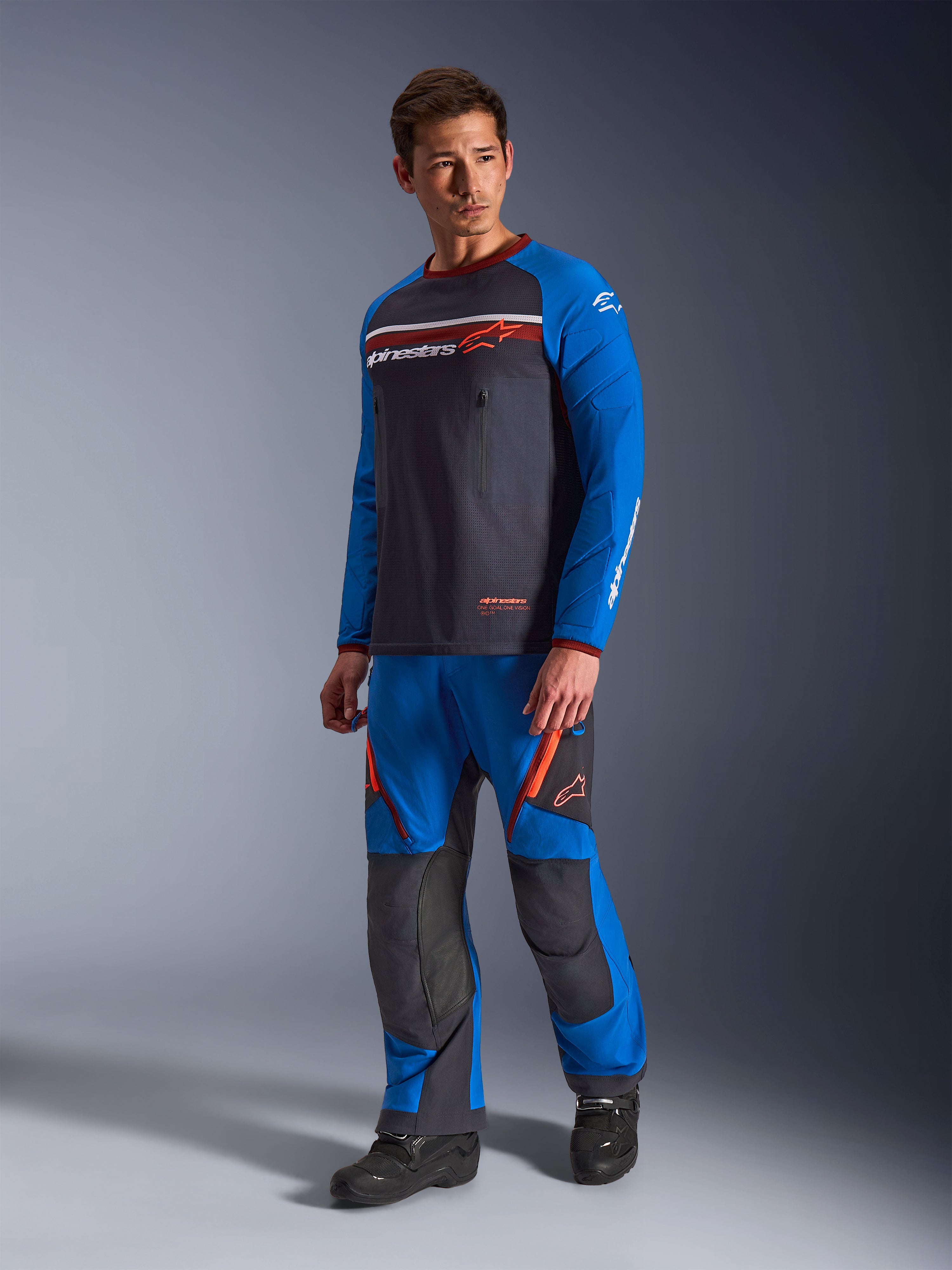 Alpinestars Maxdura Dual Jersey, grigio scuro e blu royal, maglia da moto adventure con torso in rete e maniche blu royal, indossata da un modello con pantaloni da guida off-road coordinati, dotata di tasche sul petto con cerniera e marchio Alpinestars.