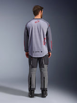 Modello uomo visto da dietro con indosso la maglia Alpinestars Maxdura Dual Jersey, maglia adventure grigio medio e grigio scuro con dettagli del logo rossi, costruzione posteriore in mesh, abbinata a pantaloni off-road grigi coordinati e stivali neri