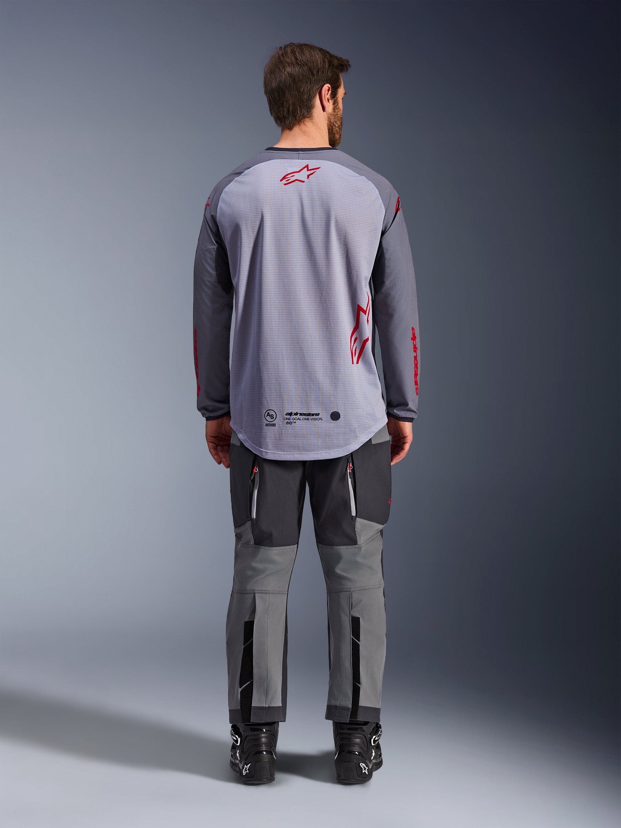 Modello uomo visto da dietro con indosso la maglia Alpinestars Maxdura Dual Jersey, maglia adventure grigio medio e grigio scuro con dettagli del logo rossi, costruzione posteriore in mesh, abbinata a pantaloni off-road grigi coordinati e stivali neri