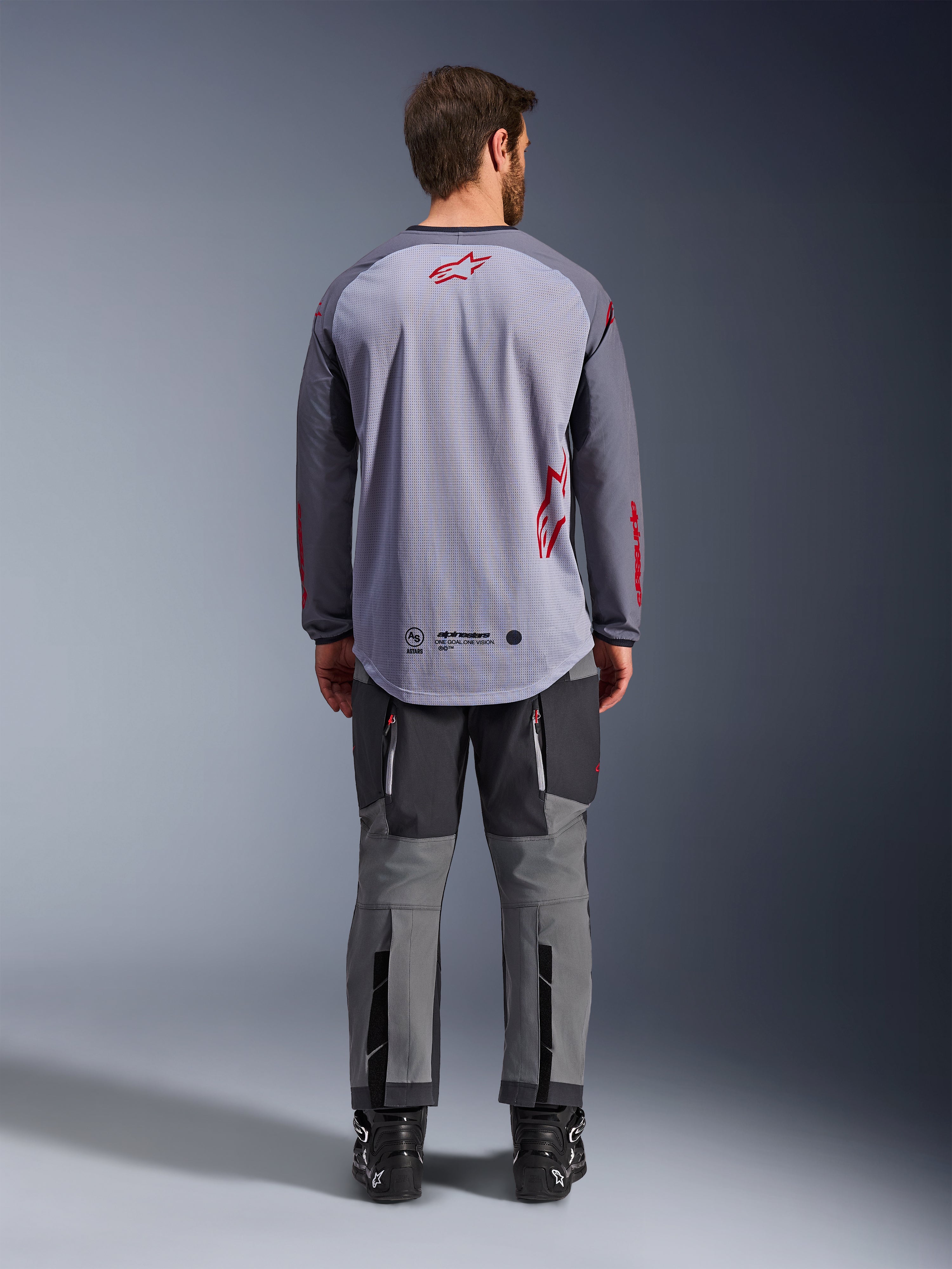 Modello uomo visto da dietro con indosso la maglia Alpinestars Maxdura Dual Jersey, maglia adventure grigio medio e grigio scuro con dettagli del logo rossi, costruzione posteriore in mesh, abbinata a pantaloni off-road grigi coordinati e stivali neri