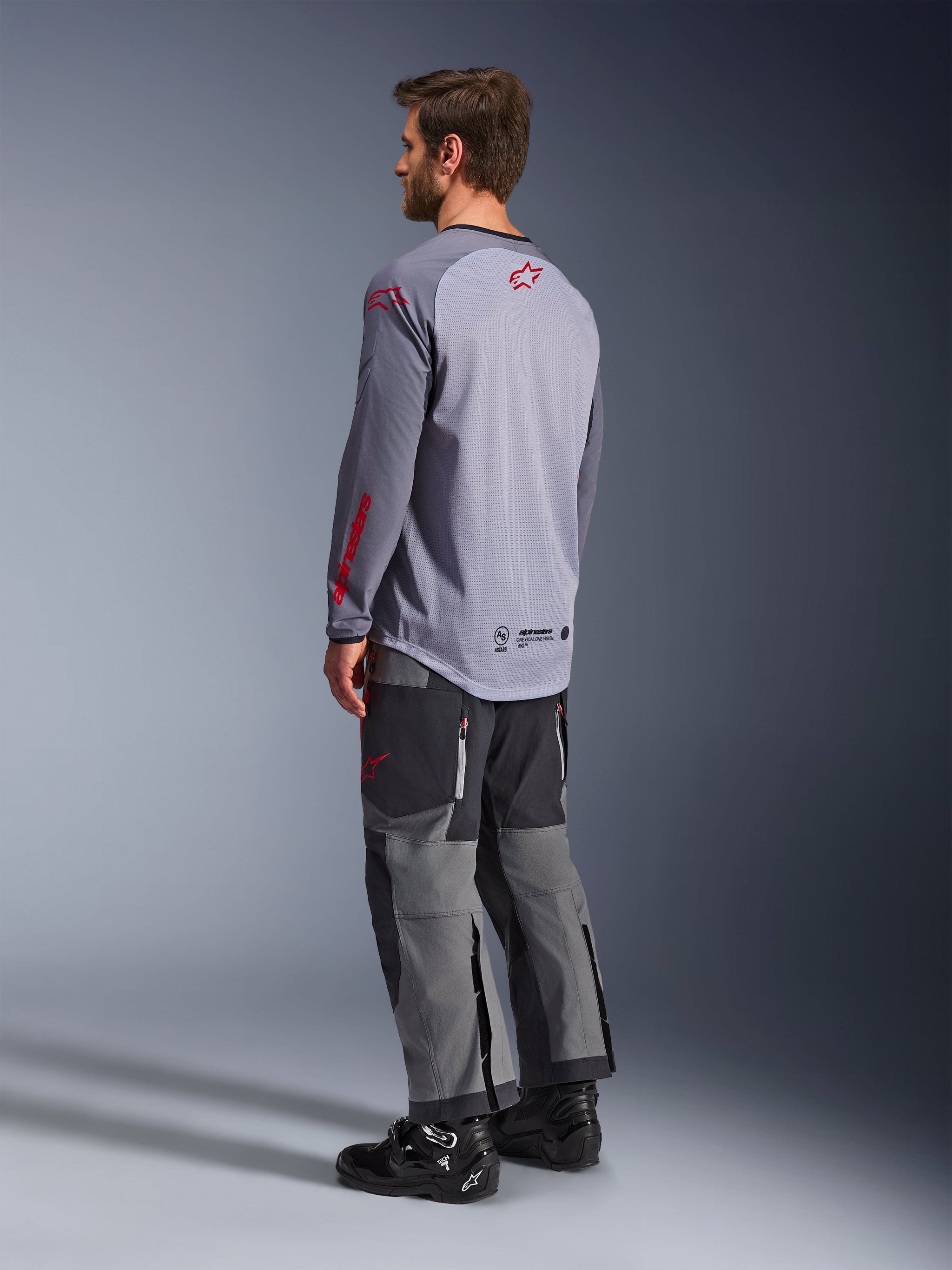 Alpinestars Maxdura Dual Jersey, maglia tecnica grigio chiaro con loghi Alpinestars rossi, vista posteriore con torso in rete e maniche lunghe, abbinata a pantaloni tecnici antracite e stivali da moto neri