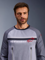 Alpinestars Maxdura Dual Jersey, grigio medio e grigio scuro, indossata da un modello, con busto in rete per la ventilazione, doppie tasche sul petto con cerniera, branding 'alpinestars' bianco e logo a stella rosso, abbigliamento tecnico per avventura fuoristrada