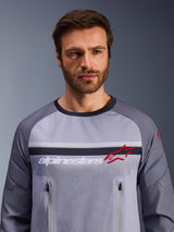 Alpinestars Maxdura Dual Jersey, grigio medio e grigio scuro, indossata da un modello, con busto in rete per la ventilazione, doppie tasche sul petto con cerniera, branding 'alpinestars' bianco e logo a stella rosso, abbigliamento tecnico per avventura fuoristrada