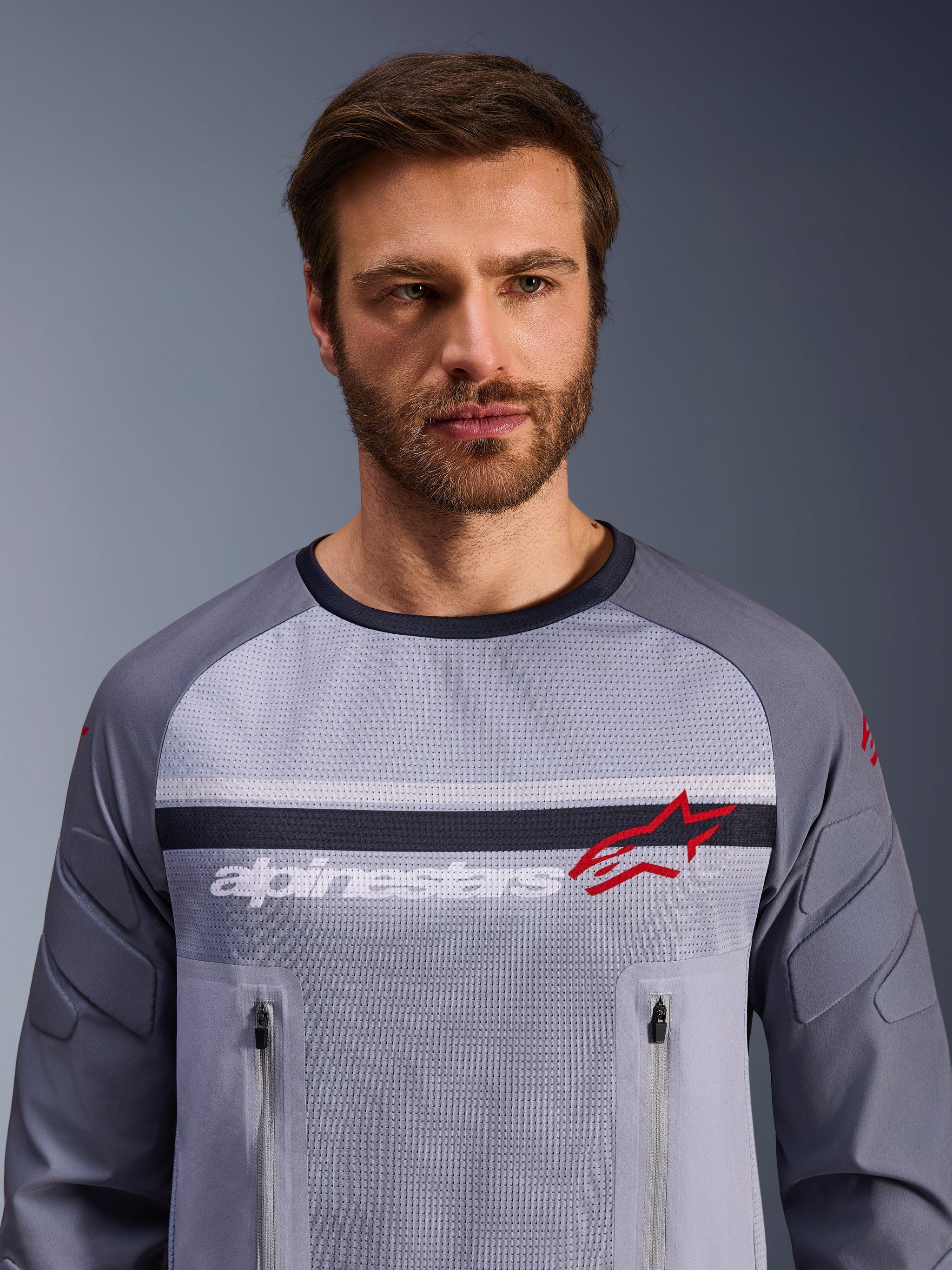 Alpinestars Maxdura Dual Jersey, grigio medio e grigio scuro, indossata da un modello, con busto in rete per la ventilazione, doppie tasche sul petto con cerniera, branding 'alpinestars' bianco e logo a stella rosso, abbigliamento tecnico per avventura fuoristrada