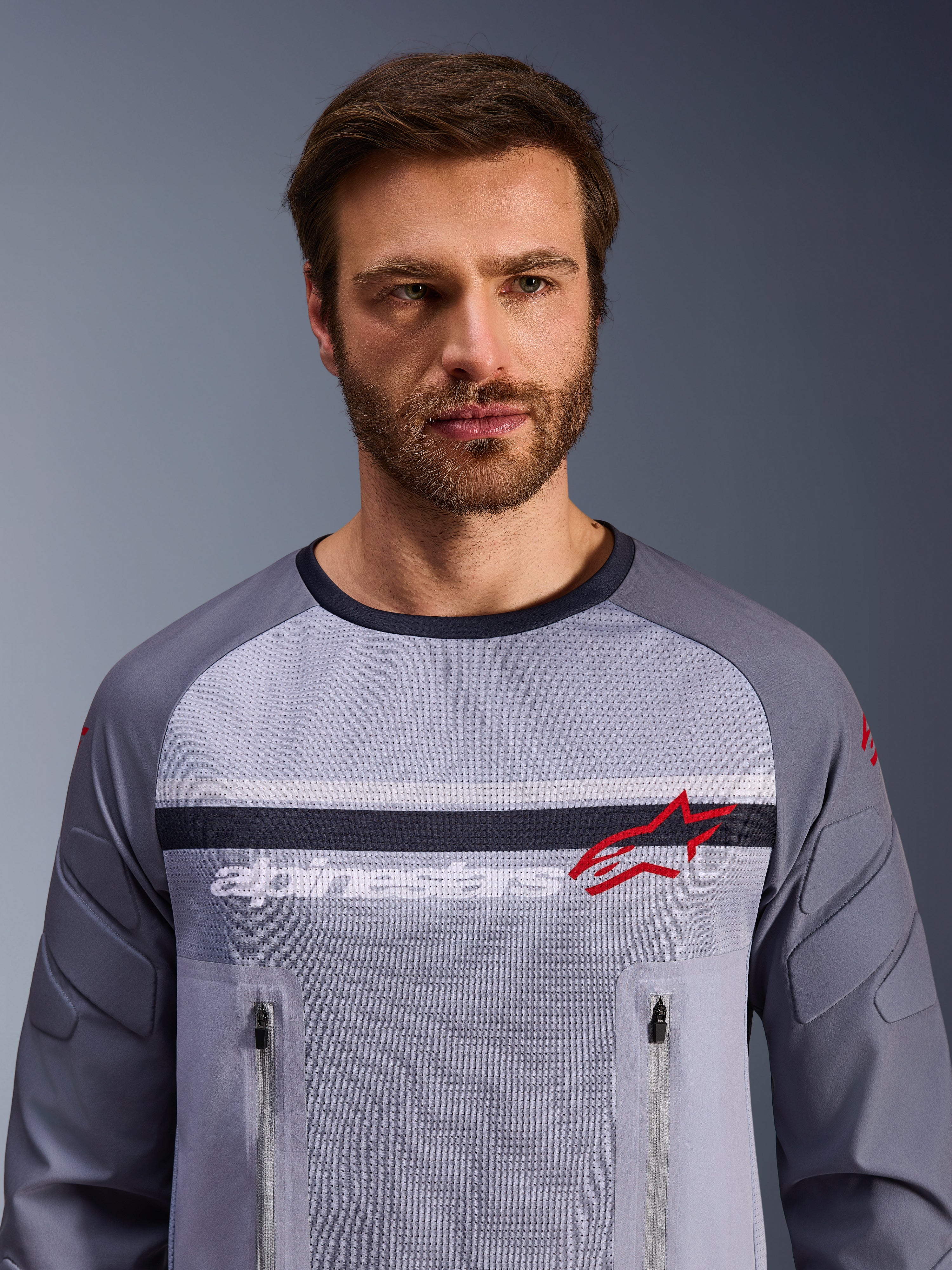Alpinestars Maxdura Dual Jersey, grigio medio e grigio scuro, indossata da un modello, con busto in rete per la ventilazione, doppie tasche sul petto con cerniera, branding 'alpinestars' bianco e logo a stella rosso, abbigliamento tecnico per avventura fuoristrada