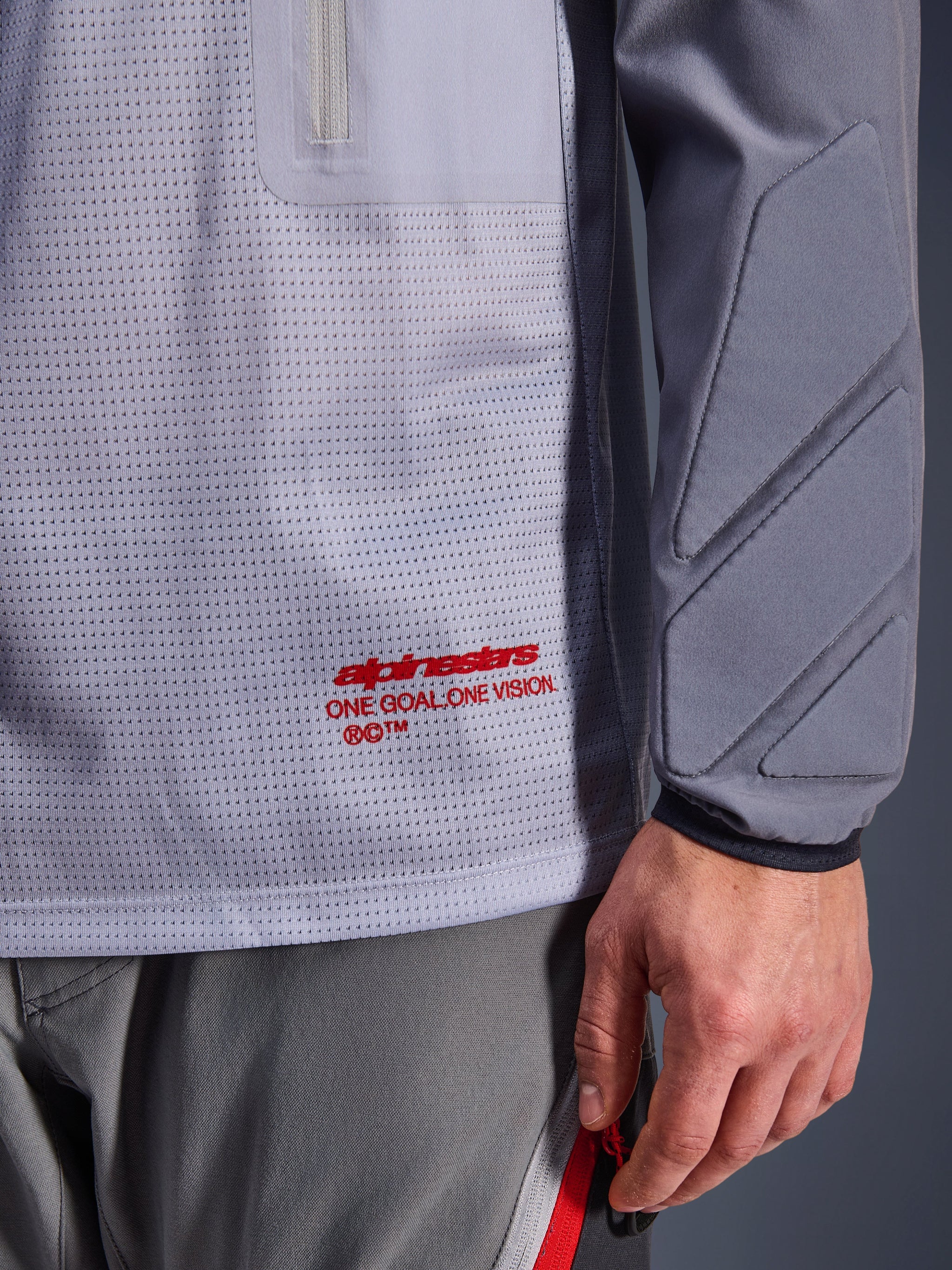 Alpinestars Maxdura Dual Jersey, Grigio Medio e Grigio Scuro, primo piano che mostra il torso in rete traspirante con marchio rosso, dettaglio della tasca con cerniera e imbottitura integrata sulla manica, abbigliamento tecnico da moto adventure