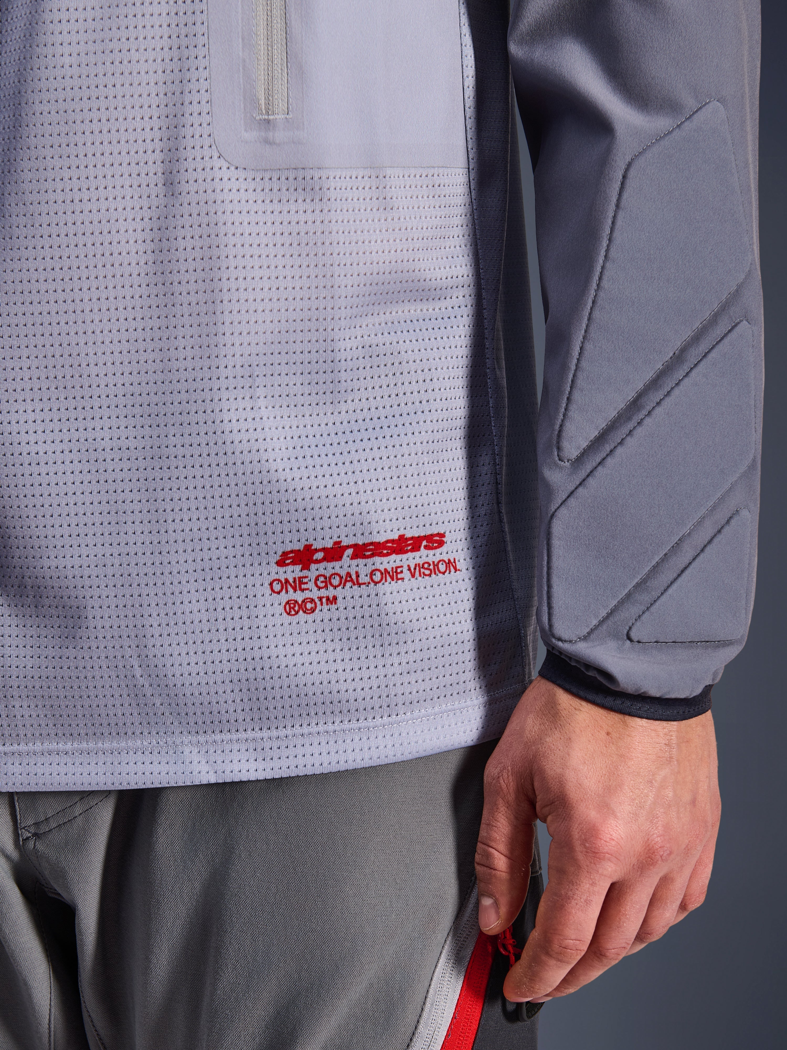 Alpinestars Maxdura Dual Jersey, Grigio Medio e Grigio Scuro, primo piano che mostra il torso in rete traspirante con marchio rosso, dettaglio della tasca con cerniera e imbottitura integrata sulla manica, abbigliamento tecnico da moto adventure