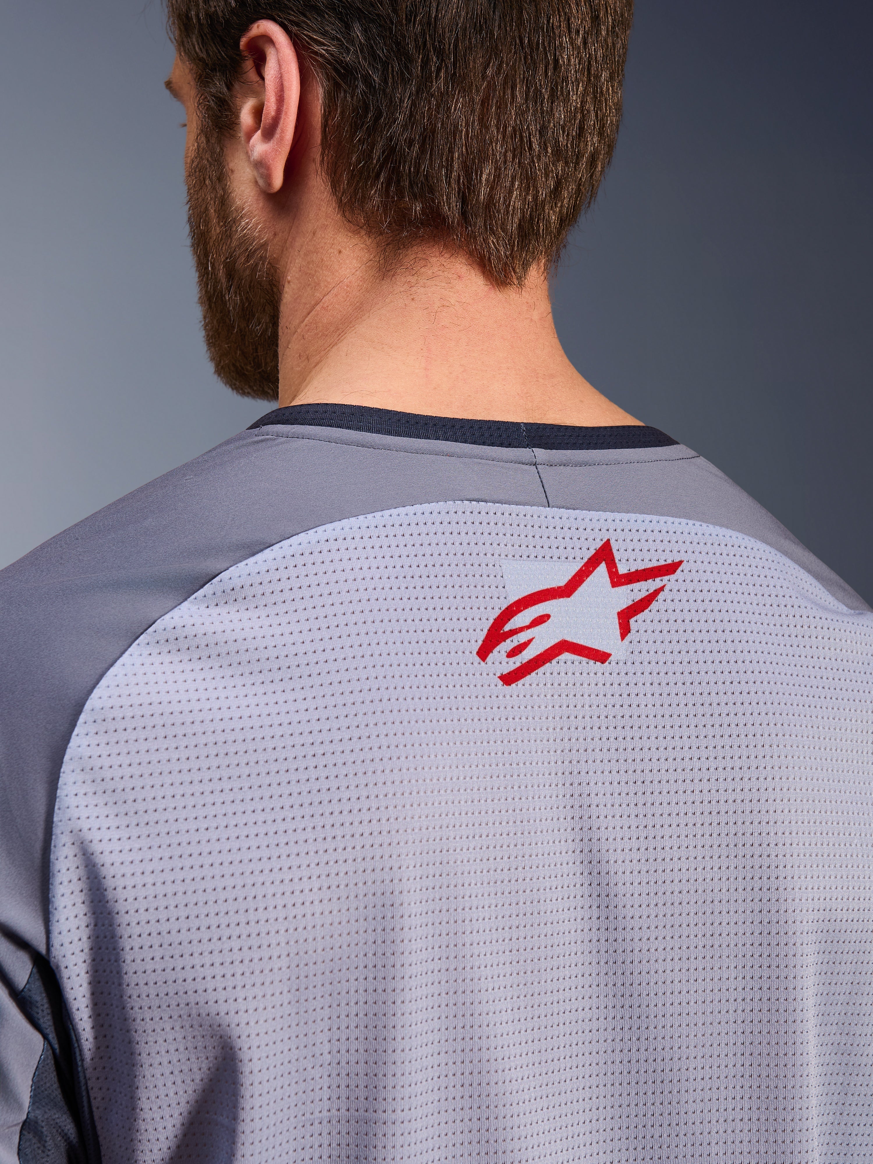 Alpinestars Maxdura Dual Jersey in Grigio Medio e Grigio Scuro, primo piano della parte superiore della schiena e del colletto, piccolo logo Alpinestars a stella rosso su tessuto a rete traspirante grigio chiaro, bordo del collo grigio scuro, progettata per la guida adventure off-road