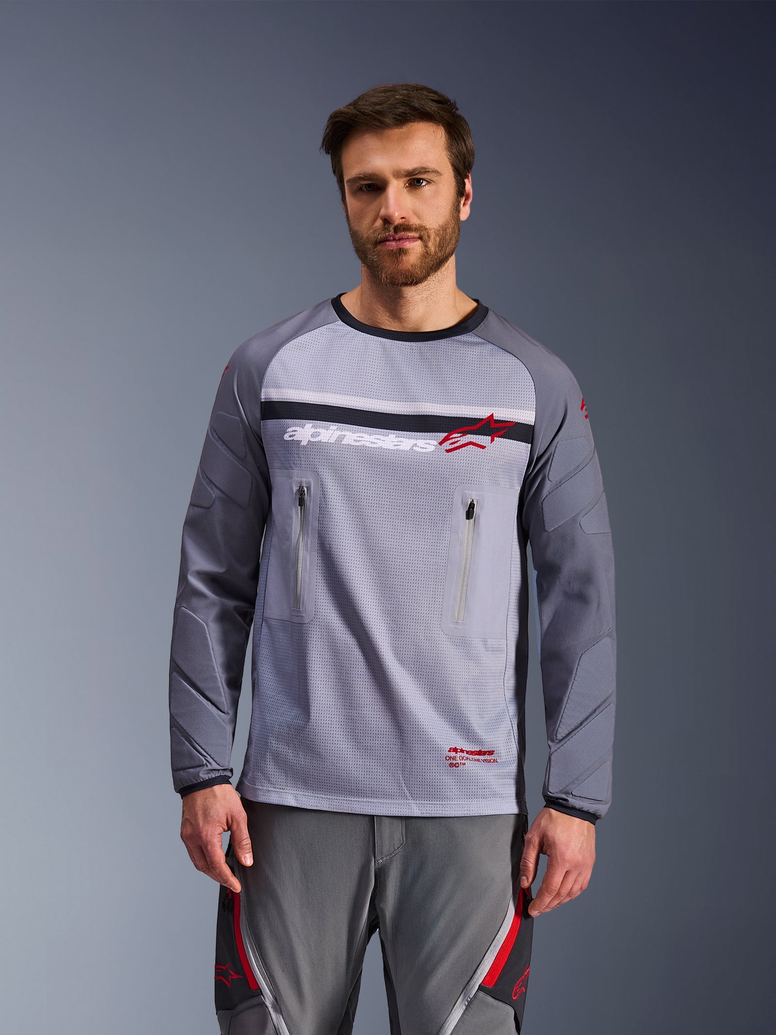 Alpinestars Maxdura Dual Jersey, grigio medio e grigio scuro, indossata da un modello con pantaloni grigi coordinati, dotata di tasche sul petto con cerniera, costruzione del busto in rete e maniche imbottite resistenti all'abrasione, abbigliamento adventure moto off-road
