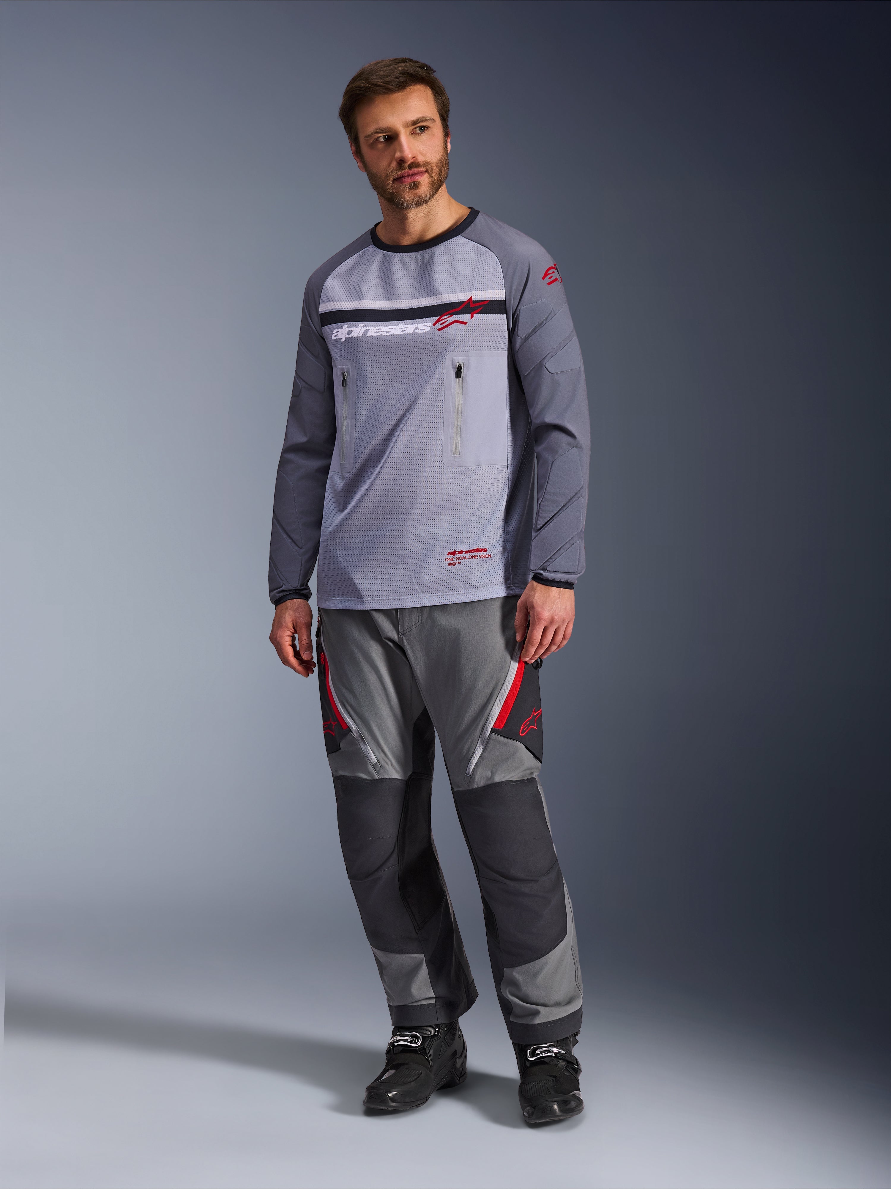 Un uomo che indossa la Alpinestars Maxdura Dual Jersey in Grigio Medio e Grigio Scuro, abbinata a pantaloni adventure coordinati grigi e neri e stivali neri, caratterizzata da un torso in rete traspirante, tasche sul petto con cerniera e dettagli del logo rossi.