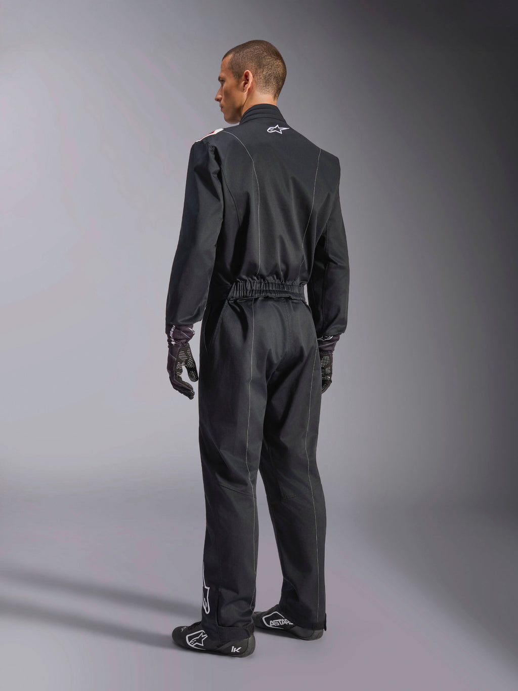 Alpinestars Kart Indoor V2 Suit, tuta da kart intera nera mostrata da un'angolazione posteriore di tre quarti, con vita elastica e logo bianco sul collo, indossata con guanti e scarpe da corsa neri, design resistente per il karting ricreativo