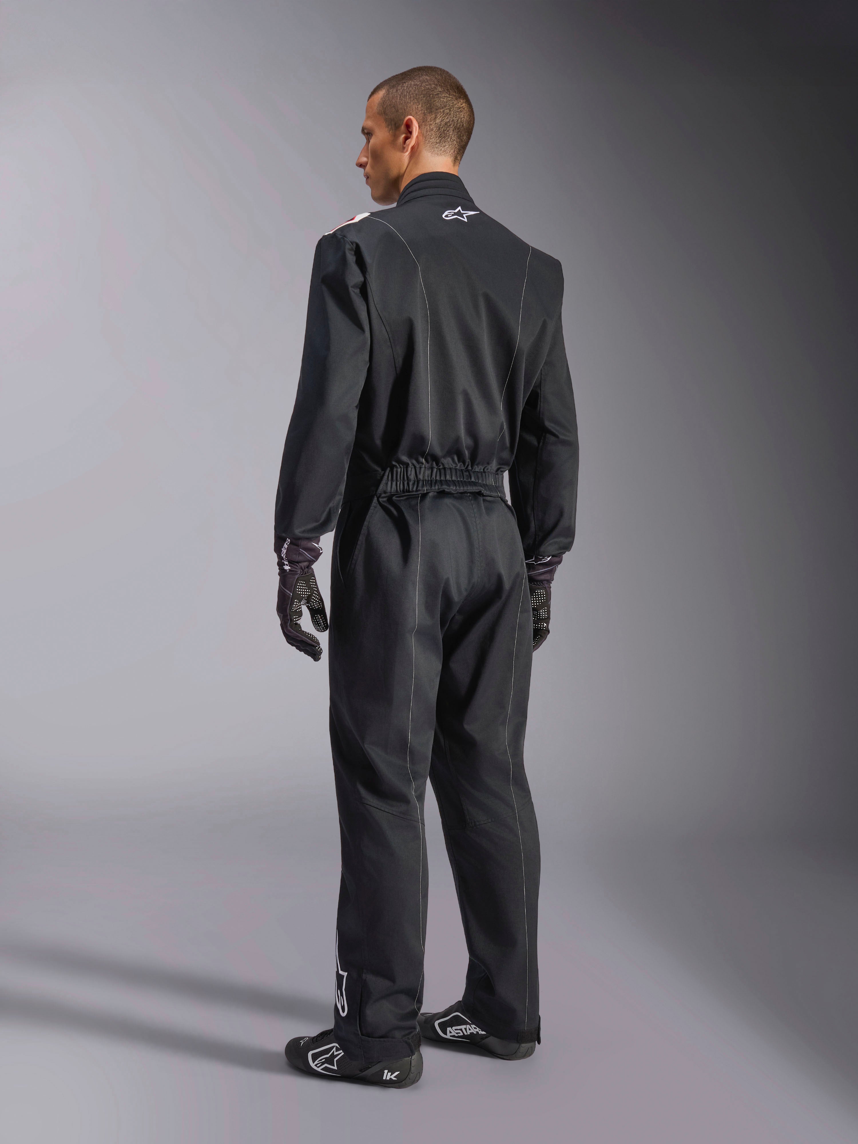 Alpinestars Kart Indoor V2 Suit, tuta da kart intera nera mostrata da un'angolazione posteriore di tre quarti, con vita elastica e logo bianco sul collo, indossata con guanti e scarpe da corsa neri, design resistente per il karting ricreativo