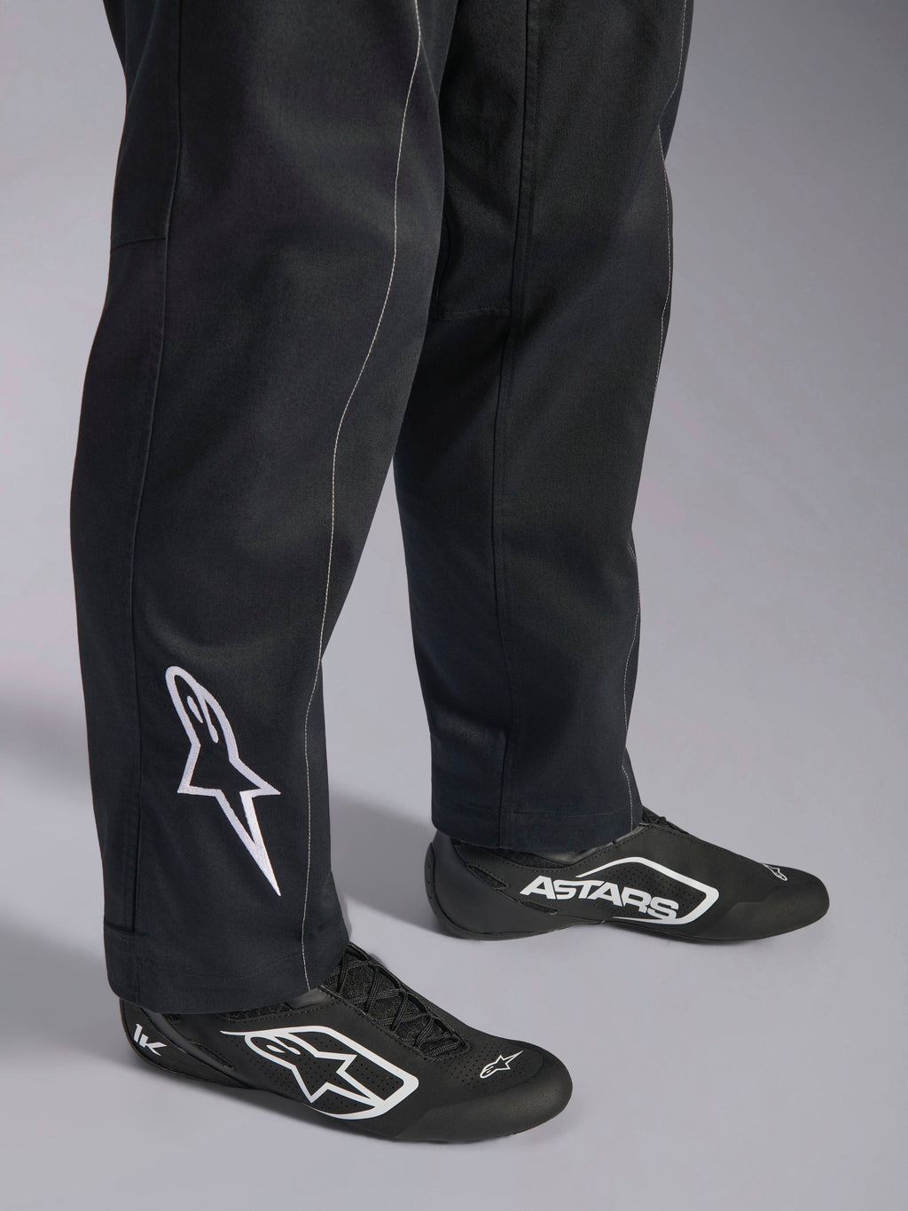 Alpinestars Kart Indoor V2 Suit, tuta da kart nera, primo piano della parte inferiore delle gambe e dei piedi con logo Alpinestars bianco sul polpaccio, abbinata a scarpe da corsa bianche e nere coordinate, progettata per il karting ricreativo.