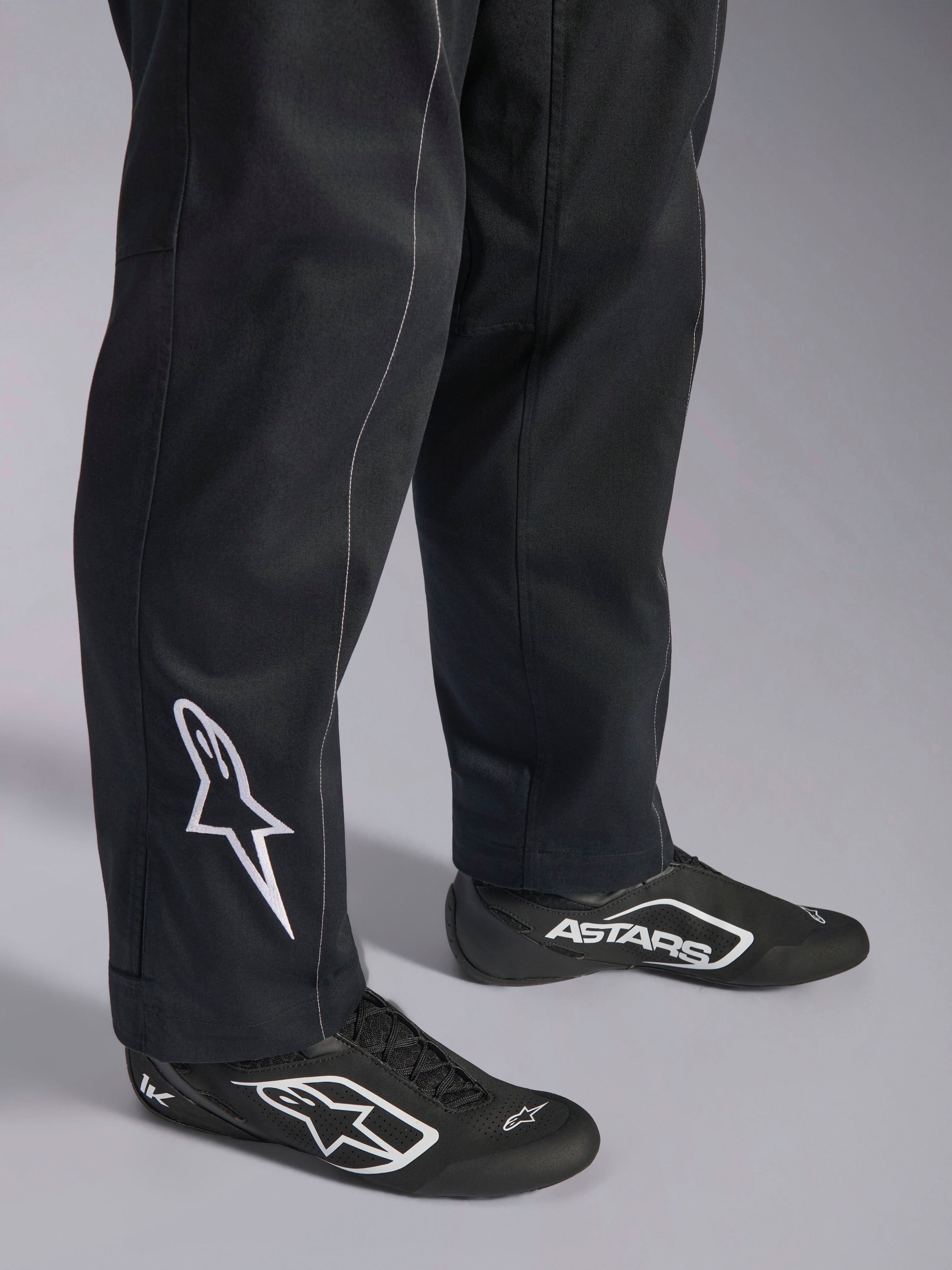 Alpinestars Kart Indoor V2 Suit, tuta da kart nera, primo piano della parte inferiore delle gambe e dei piedi con logo Alpinestars bianco sul polpaccio, abbinata a scarpe da corsa bianche e nere coordinate, progettata per il karting ricreativo.