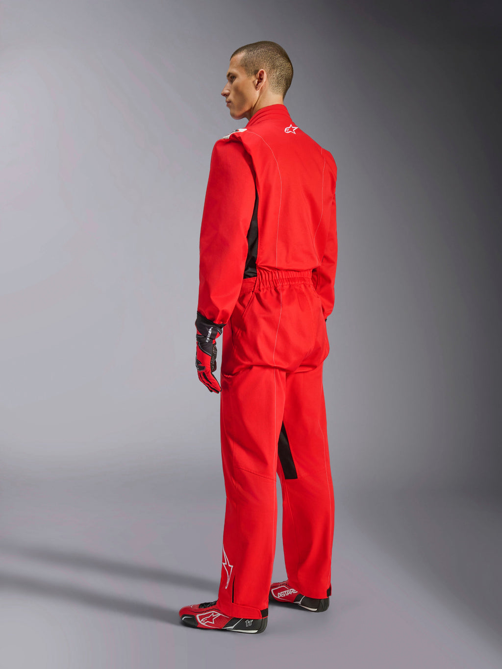 Alpinestars Kart Indoor V2 Suit, tuta da kart rossa, vista posteriore di tre quarti su un modello, indossata con guanti rossi coordinati e scarpe da corsa, costruzione in twill di policotone a strato singolo, equipaggiamento professionale da motorsport per il karting indoor.