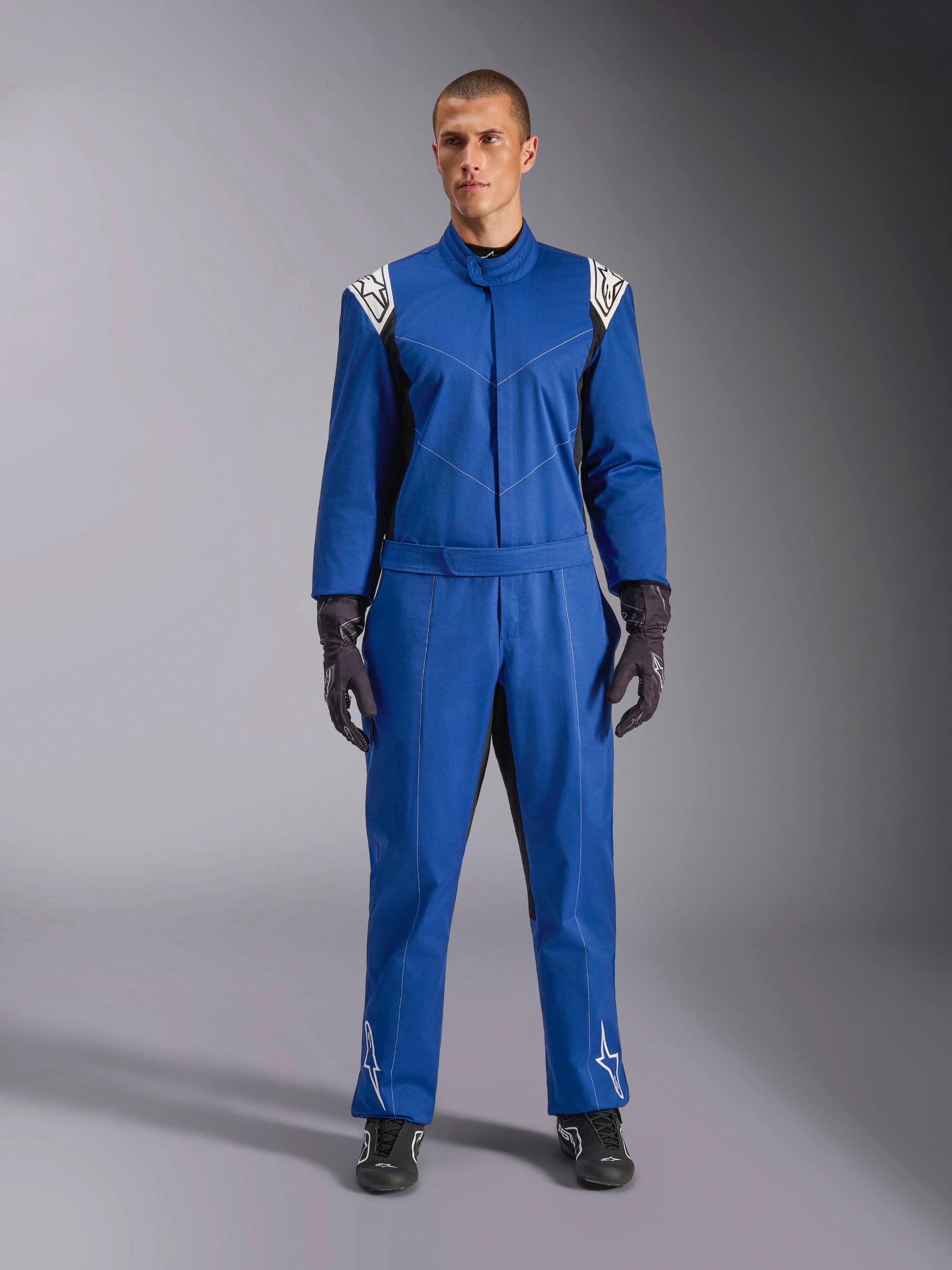 Modello che indossa la tuta Alpinestars Kart Indoor V2 in blu, tuta da kart intera con loghi a stella bianchi su spalle e stinchi, cuciture a contrasto bianche, cintura elastica in vita, abbinata a guanti da corsa e calzature neri, progettata per l'uso ricreativo in pista
