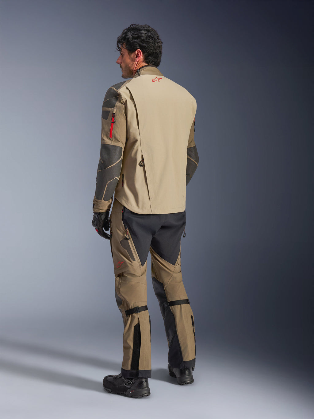Giacca Alpinestars Nazca 3L GORE-TEX PRO, giacca da moto adventure, Walnut Rock Brown, vista posteriore di un pilota che indossa un completo da moto adventure con pantaloni e stivali coordinati, con pannelli marrone chiaro e neri, logo rosso sulla parte superiore della schiena, costruzione in tessuto resistente.