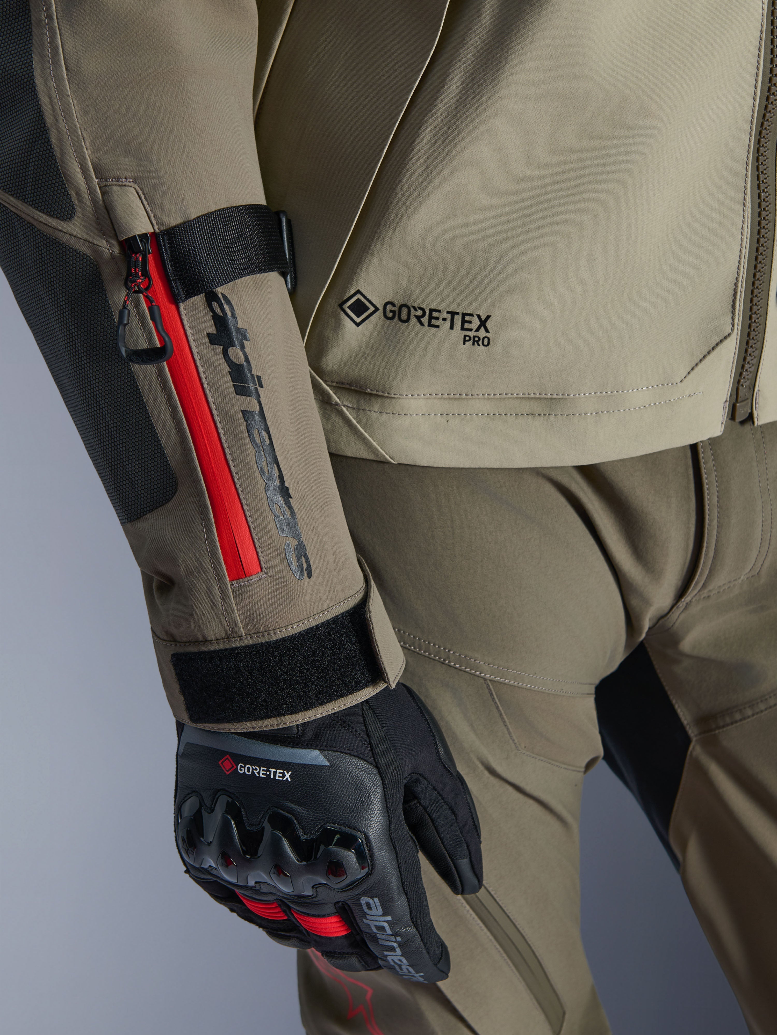 Primo piano della giacca Alpinestars Nazca 3L GORE-TEX PRO in Walnut Rock Brown, con dettagli della cerniera rossi sulla manica, logo GORE-TEX PRO e abbinata a guanti da moto adventure neri in GORE-TEX.