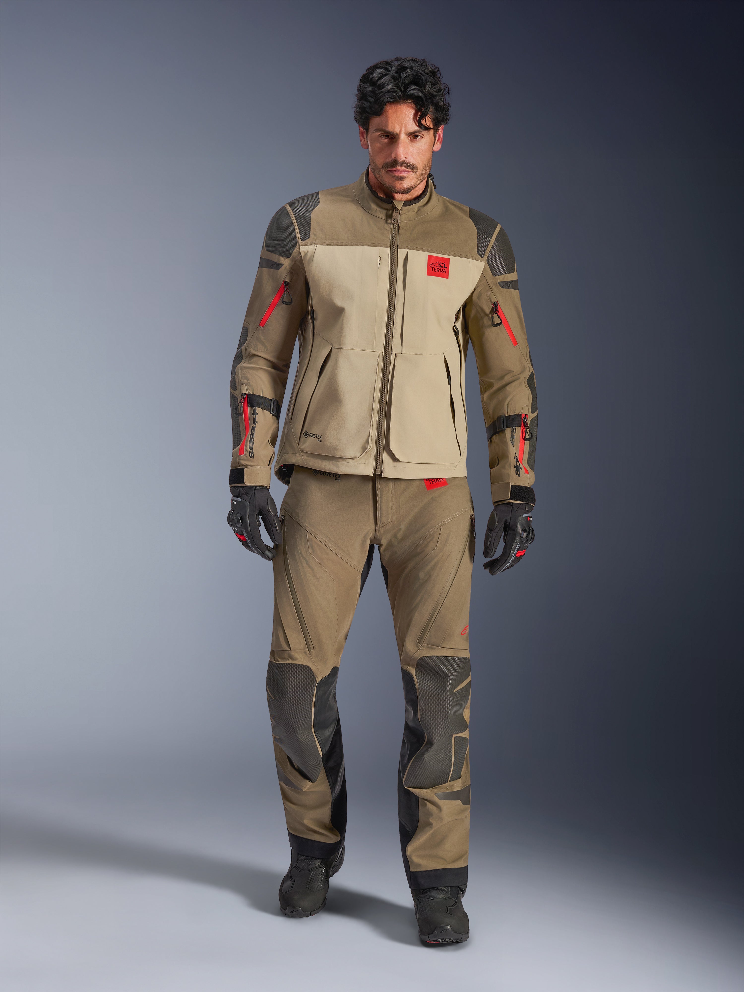 Giacca Alpinestars Nazca 3L GORE-TEX PRO, Walnut Rock Brown, giacca da moto adventure indossata con pantaloni e guanti coordinati, con dettagli delle cerniere rossi, logo 'All Terra' sul petto e costruzione in tessuto robusto.