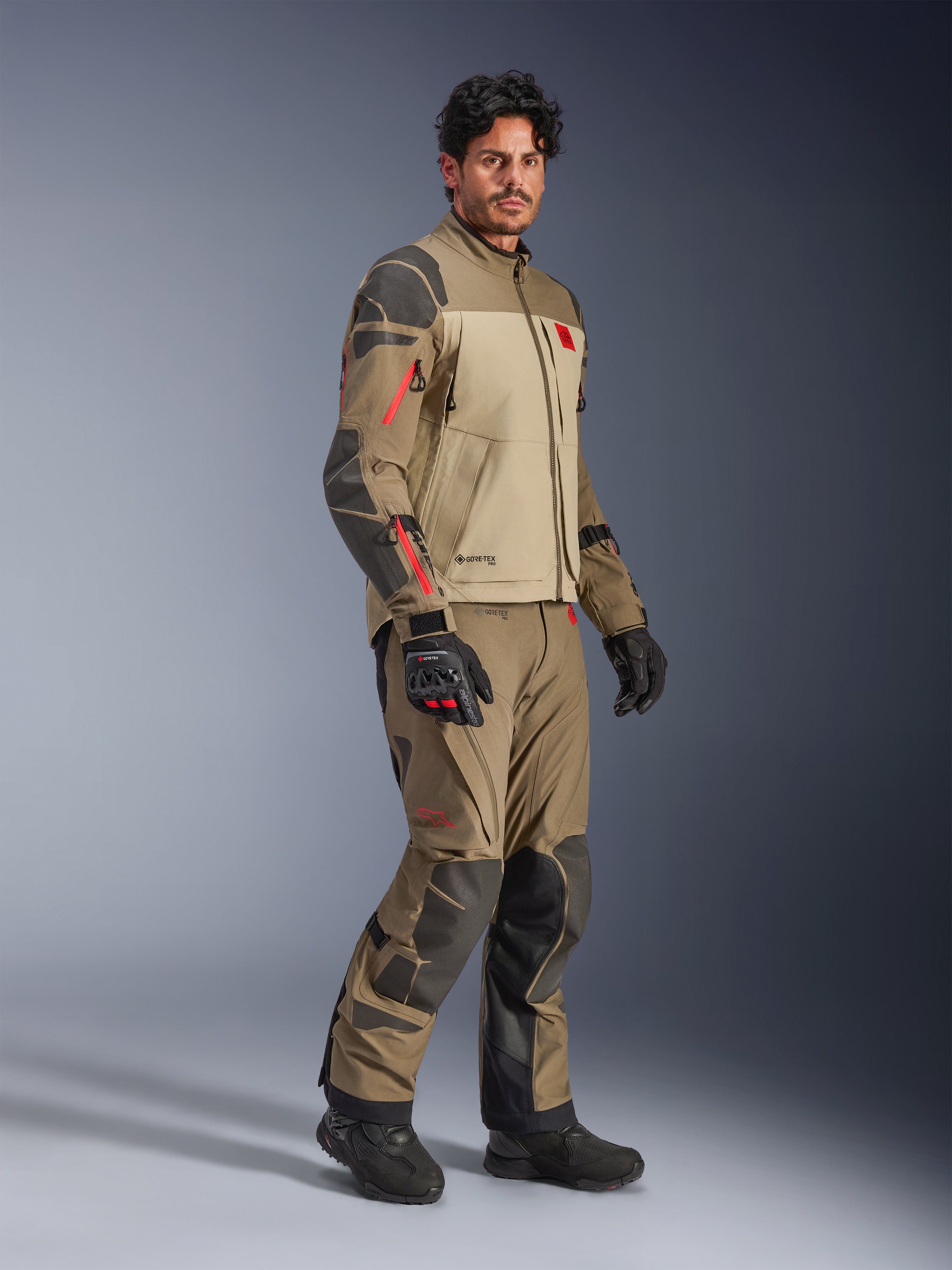 Alpinestars Nazca 3L GORE-TEX PRO Jacket, Walnut Rock Brown, giacca da moto adventure indossata con pantaloni coordinati e guanti neri, con rinforzi grigio scuro, dettagli delle cerniere rossi e guscio laminato ad alte prestazioni.