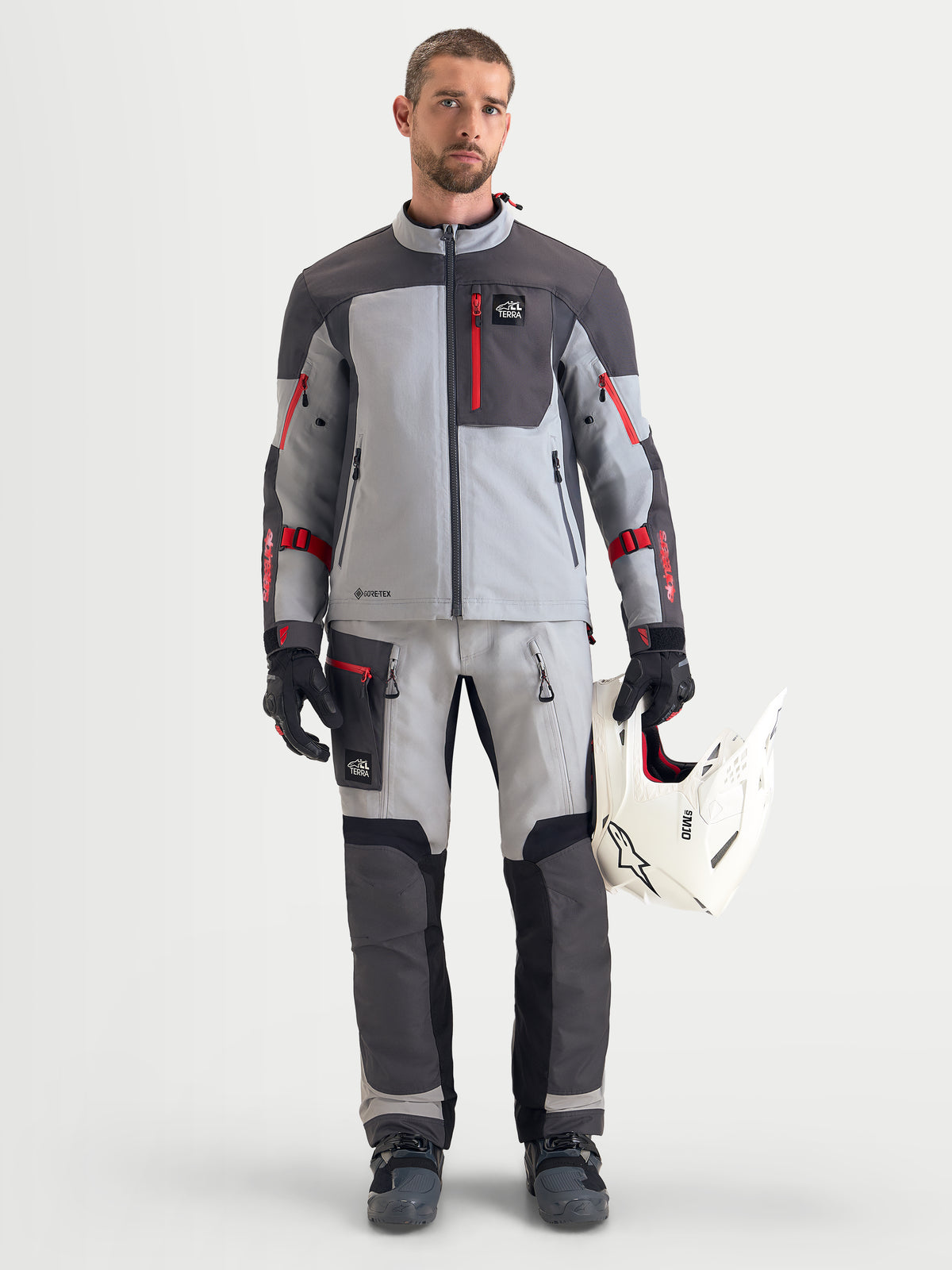 Alpinestars Arenis Gore-Tex Gear Set, completo da moto adventure da uomo in grigio e grigio scuro con dettagli rossi, vista frontale su un modello che indossa abbigliamento da guida coordinato e guanti, tenendo un casco bianco, abbigliamento impermeabile all-terrain