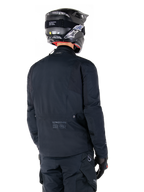 Giacca Alpinestars St-7 Prime GORE-TEX, nero, vista posteriore di un pilota che indossa la giacca con casco e pantaloni coordinati, con marchio Alpinestars e coordinate geografiche sulla parte bassa della schiena.