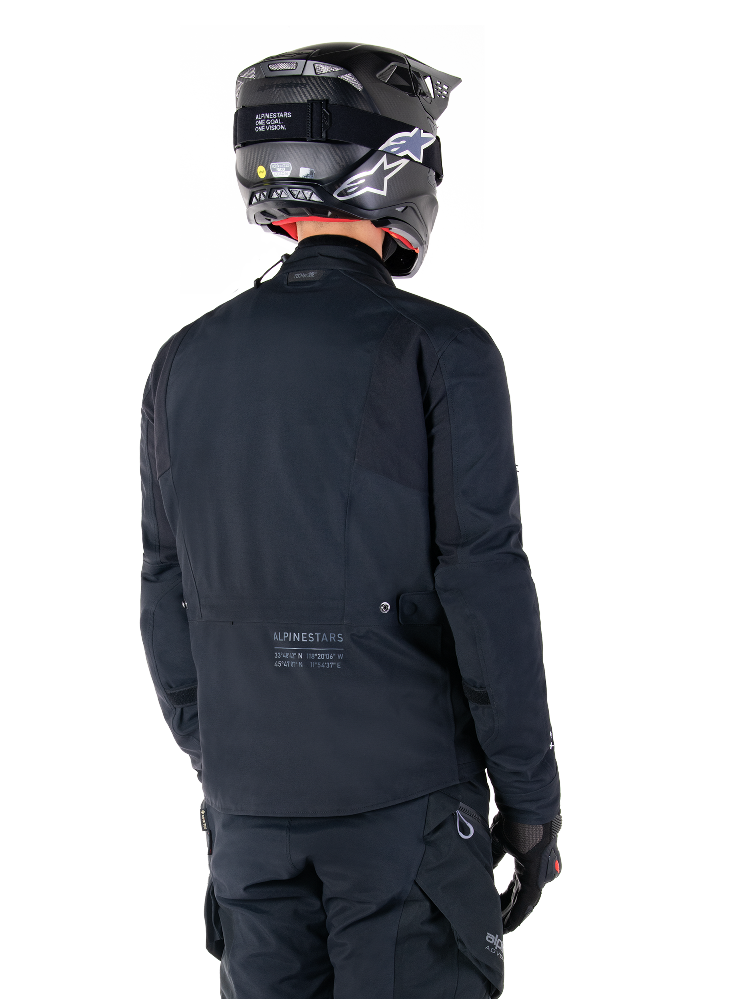Giacca Alpinestars St-7 Prime GORE-TEX, nero, vista posteriore di un pilota che indossa la giacca con casco e pantaloni coordinati, con marchio Alpinestars e coordinate geografiche sulla parte bassa della schiena.
