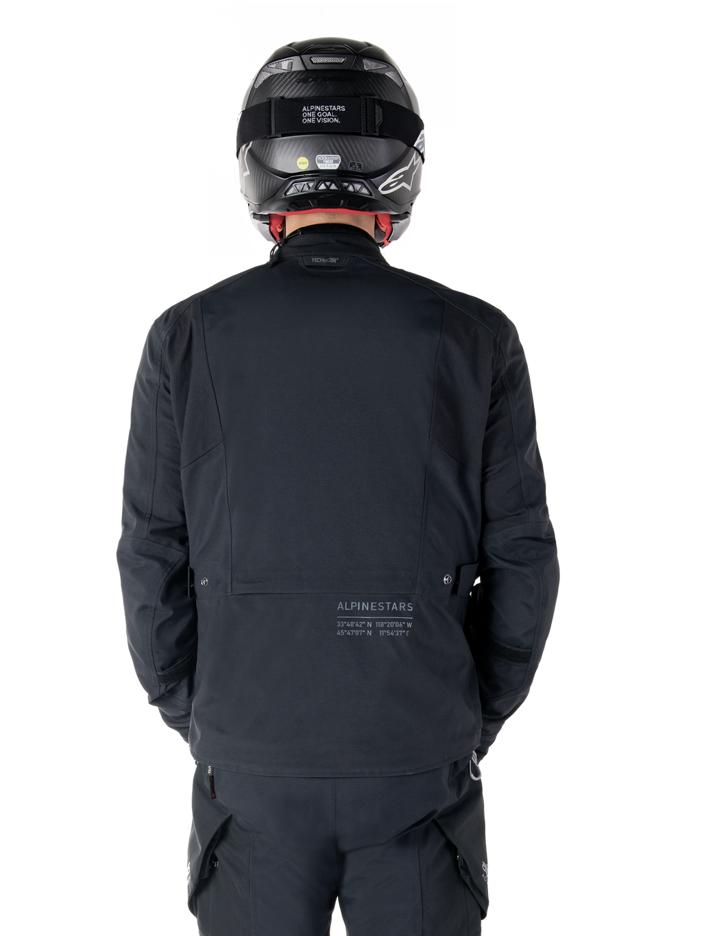 Giacca Alpinestars St-7 Prime GORE-TEX, nera, vista posteriore di un motociclista, con logo discreto e coordinate geografiche sulla parte bassa della schiena, indossata con casco e pantaloni neri coordinati, abbigliamento tecnico da motociclismo