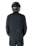 Giacca Alpinestars St-7 Prime GORE-TEX, nera, vista posteriore di un motociclista, con logo discreto e coordinate geografiche sulla parte bassa della schiena, indossata con casco e pantaloni neri coordinati, abbigliamento tecnico da motociclismo