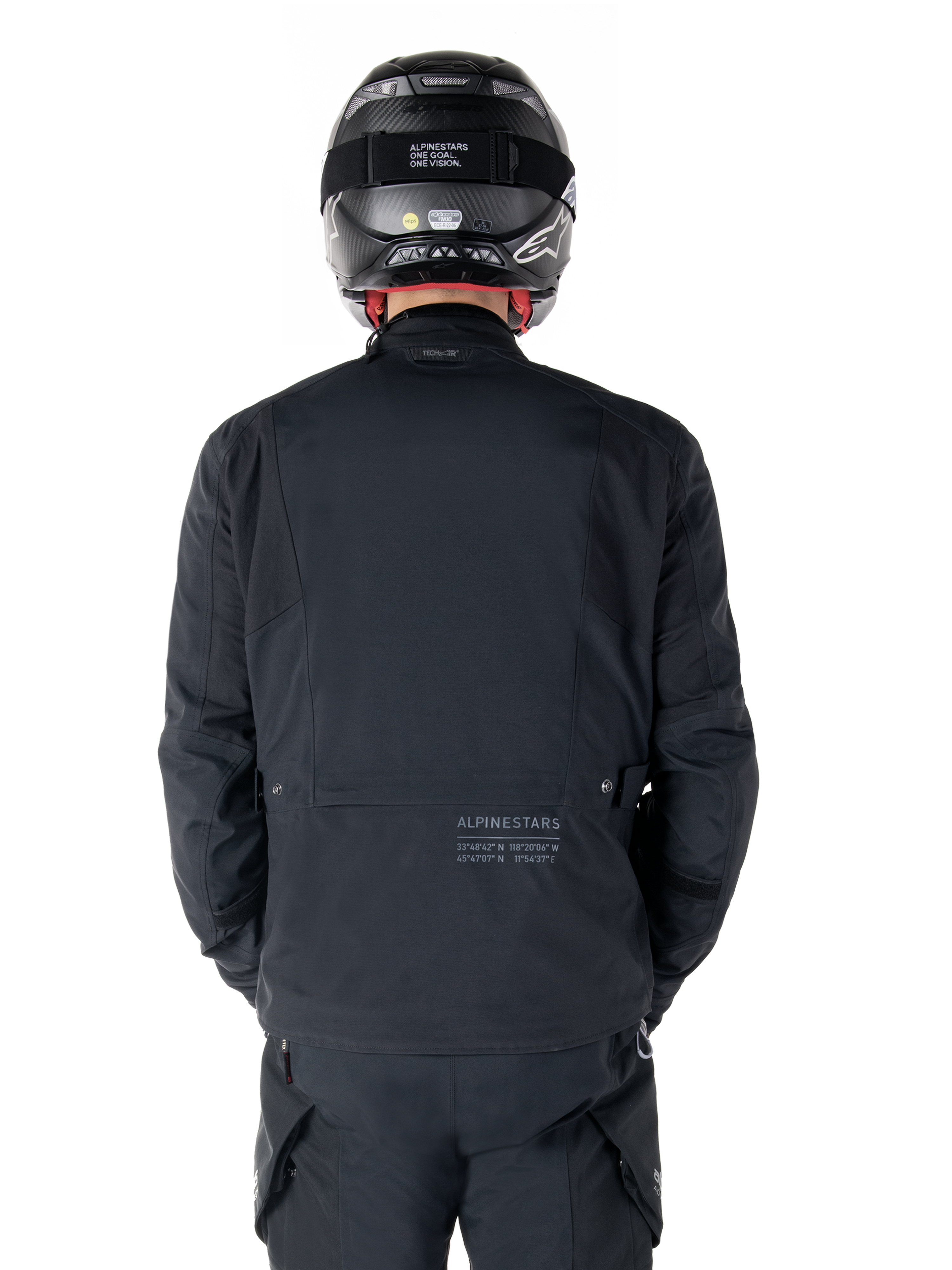 Giacca Alpinestars St-7 Prime GORE-TEX, nera, vista posteriore di un motociclista, con logo discreto e coordinate geografiche sulla parte bassa della schiena, indossata con casco e pantaloni neri coordinati, abbigliamento tecnico da motociclismo