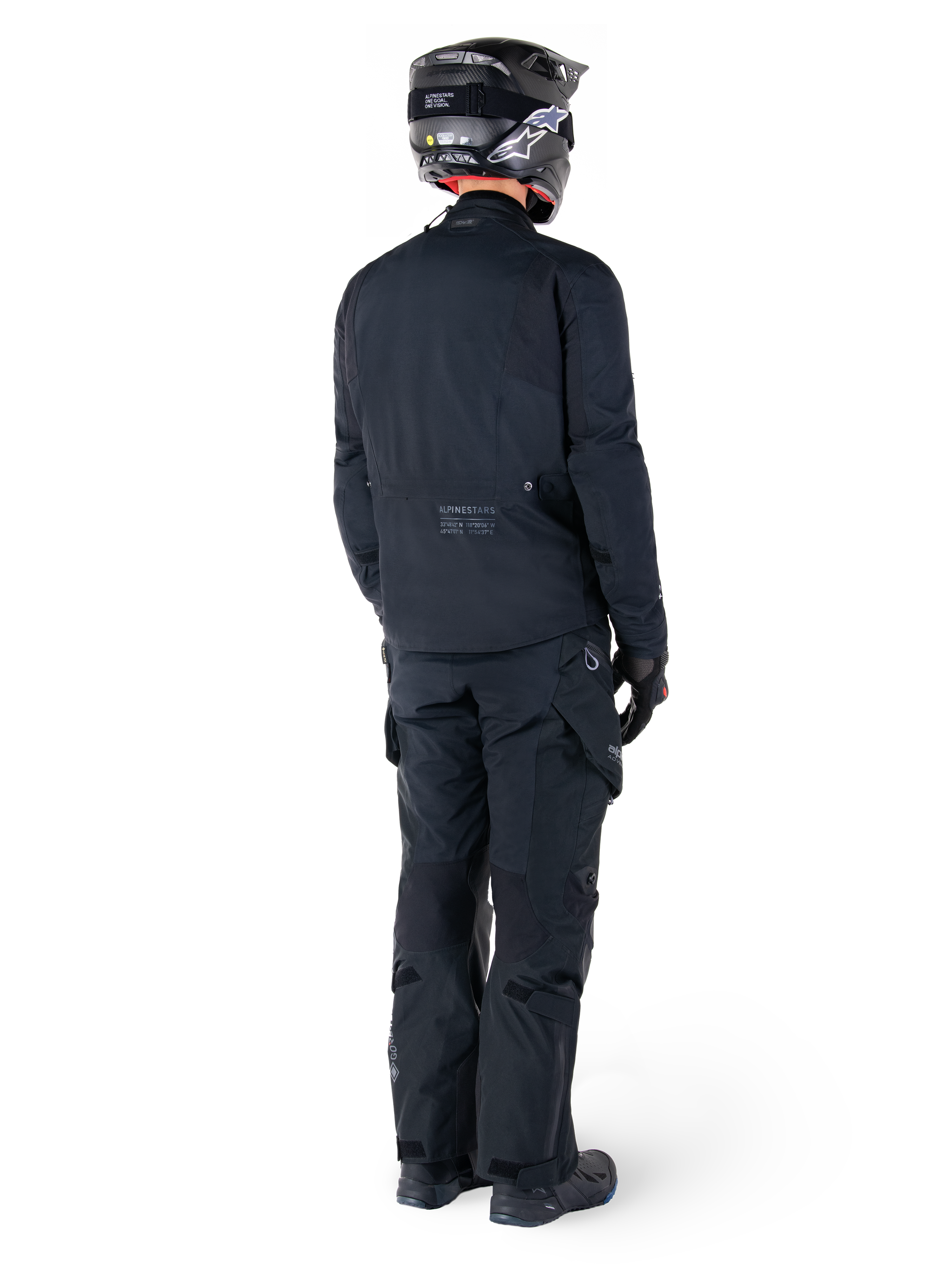 Vista posteriore di un pilota con giacca Alpinestars St-7 Prime GORE-TEX nera, pantaloni da touring neri coordinati, casco da moto nero con logo bianco e stivali neri, abbigliamento protettivo adventure completo per touring e commuting.