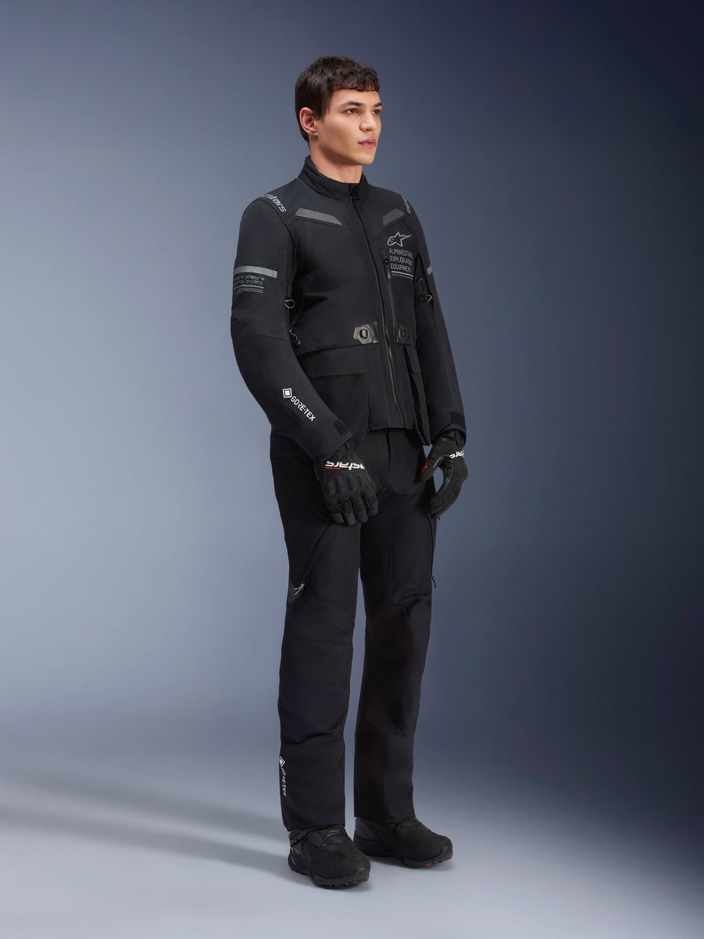 Pantaloni Alpinestars ST-7 2L GORE-TEX, indossati da un modello con la giacca ST-7 coordinata, completo da moto adventure touring total black, con logo GORE-TEX, tasche cargo, guanti e stivali neri, scatto in studio su sfondo grigio