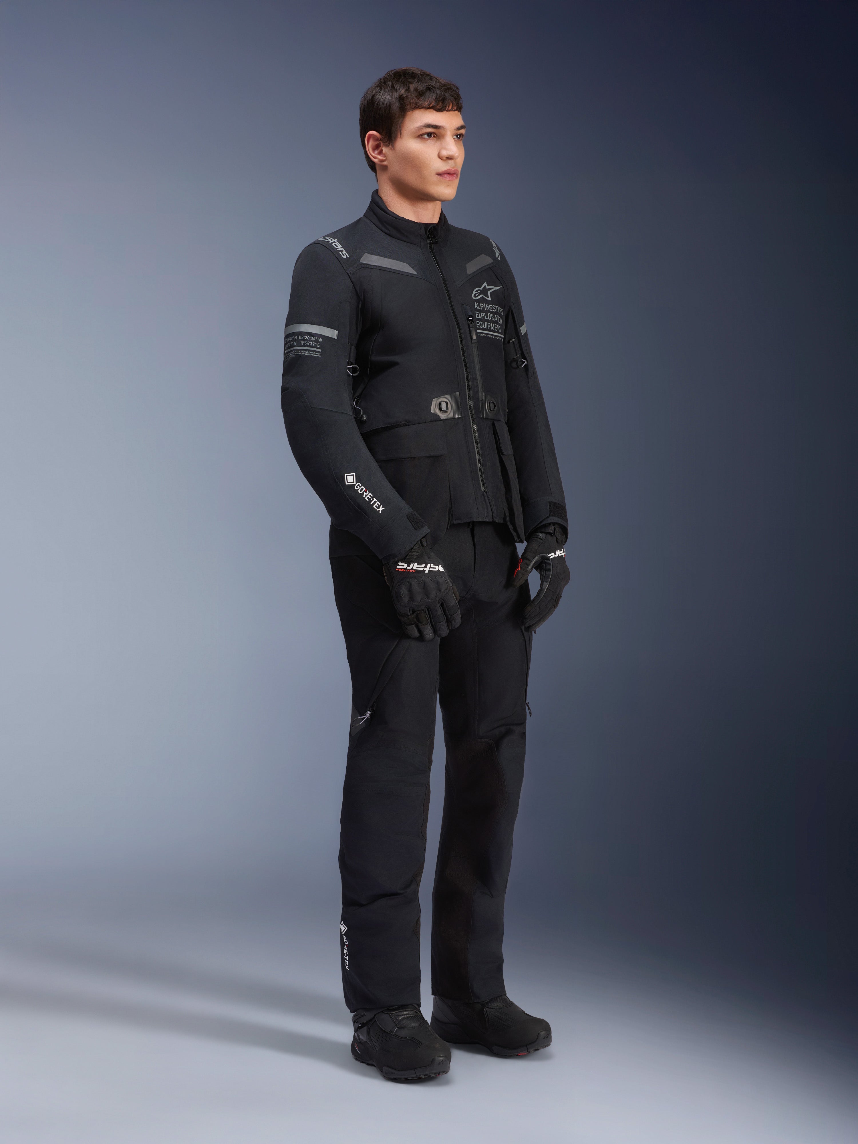 Modello con giacca Alpinestars ST-7 2L GORE-TEX in Nero Grigio Scuro, giacca da moto adventure touring, pantaloni e guanti neri coordinati, vista a figura intera di tre quarti, con logo GORE-TEX e marchio Alpinestars Exploration Equipment