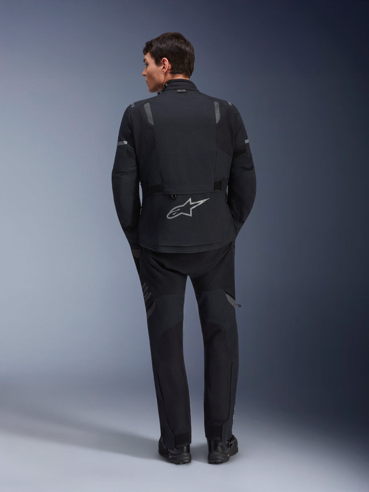 Giacca Alpinestars ST-7 2L GORE-TEX e pantaloni ST-7 2L GORE-TEX coordinati, abbigliamento da moto adventure nero e grigio scuro, vista posteriore che mostra il logo Alpinestars riflettente sulla giacca e la costruzione in tessuto tecnico.