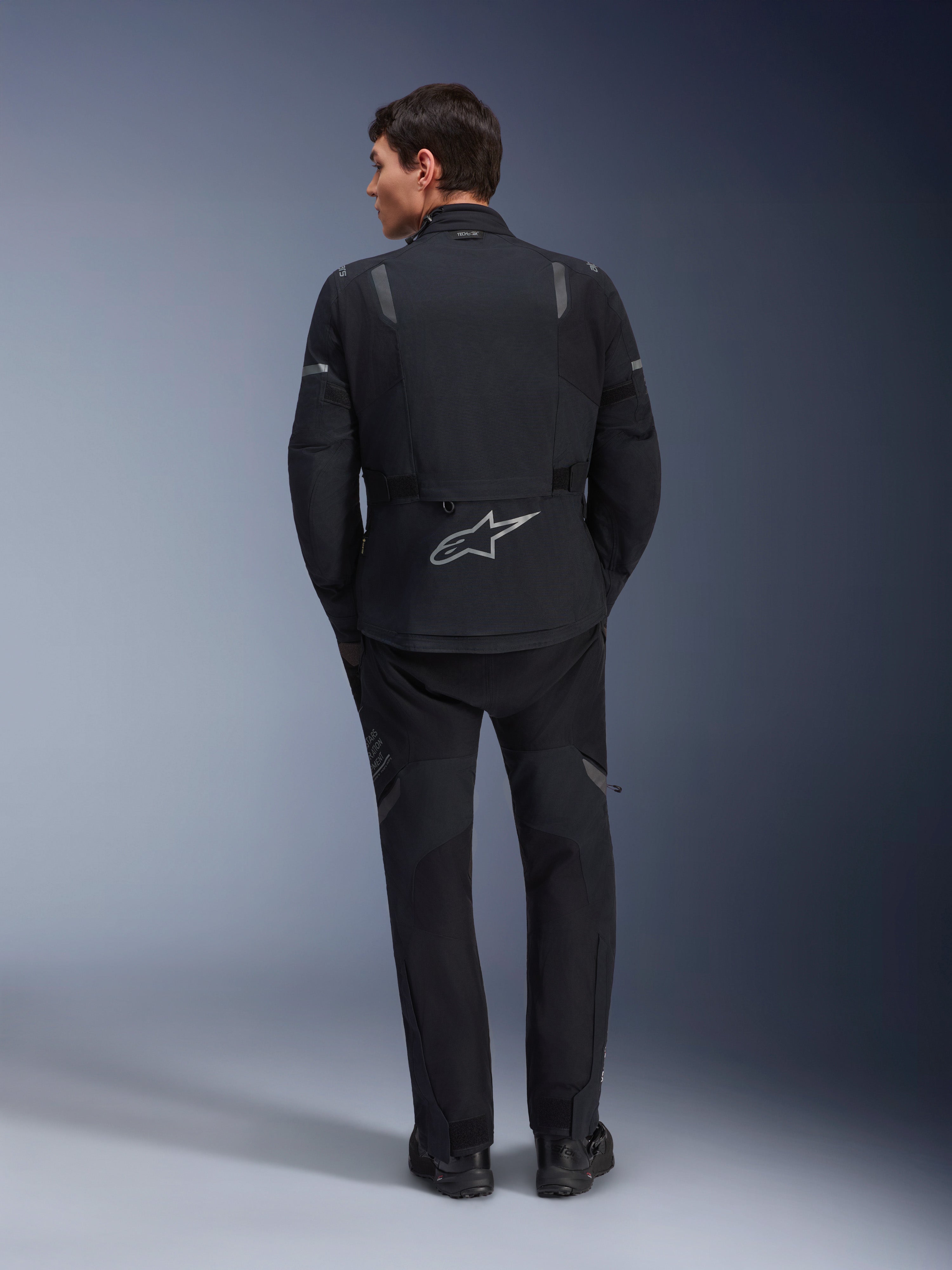 Giacca Alpinestars ST-7 2L GORE-TEX e pantaloni ST-7 2L GORE-TEX coordinati, abbigliamento da moto adventure nero e grigio scuro, vista posteriore che mostra il logo Alpinestars riflettente sulla giacca e la costruzione in tessuto tecnico.