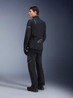 Modello che indossa la giacca Alpinestars ST-7 2L GORE-TEX in Black Dark Gray, vista posteriore di tre quarti con dettagli riflettenti e logo argento sulla parte bassa della schiena, abbigliamento da moto adventure touring abbinato a pantaloni e stivali neri.