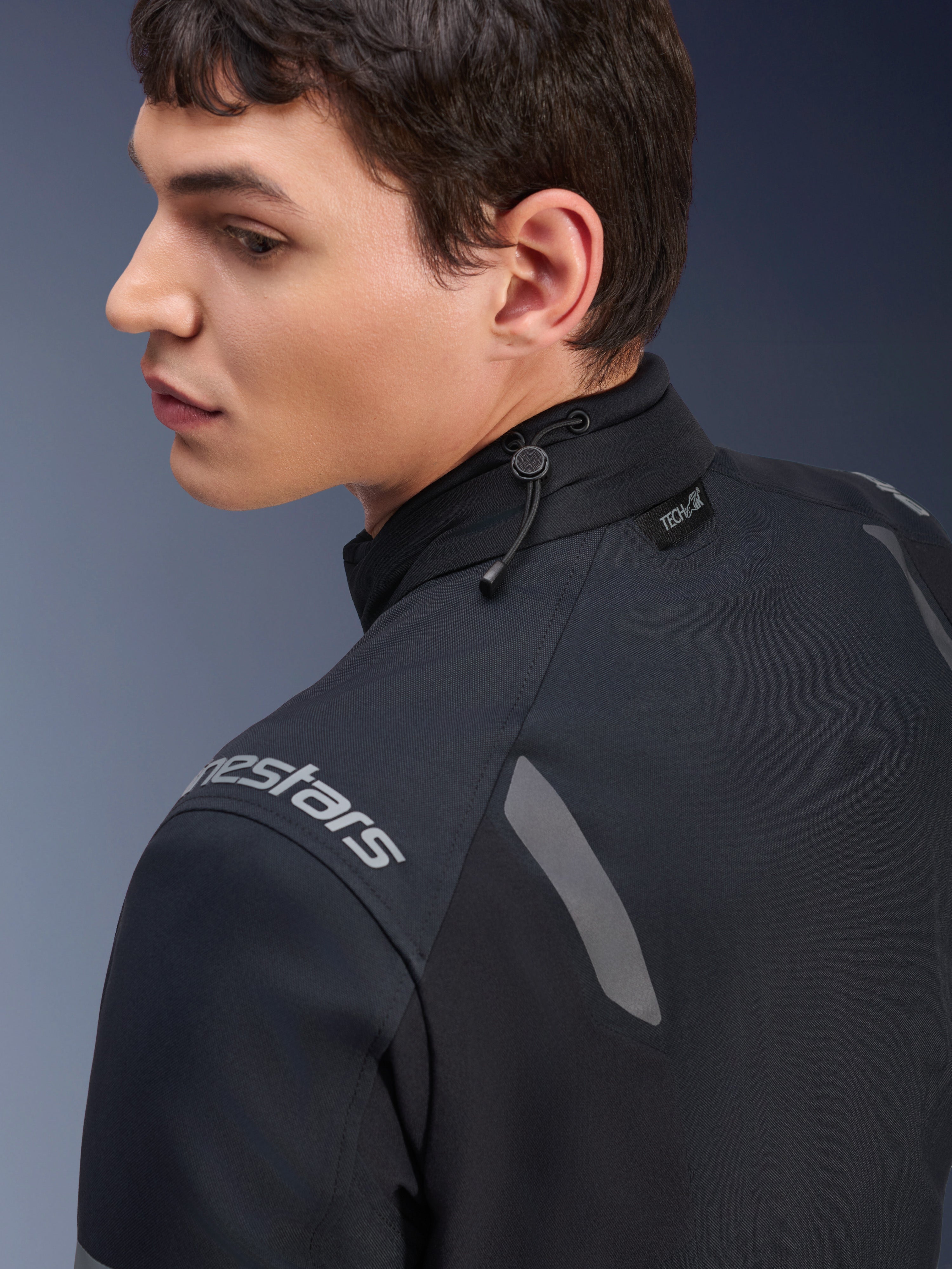 Giacca Alpinestars ST-7 2L GORE-TEX, giacca da moto nera e grigio scuro, vista ravvicinata del colletto con regolazione a cordino elastico ed etichetta Tech-Air, con dettagli riflettenti grigi e logo del brand sulla spalla.