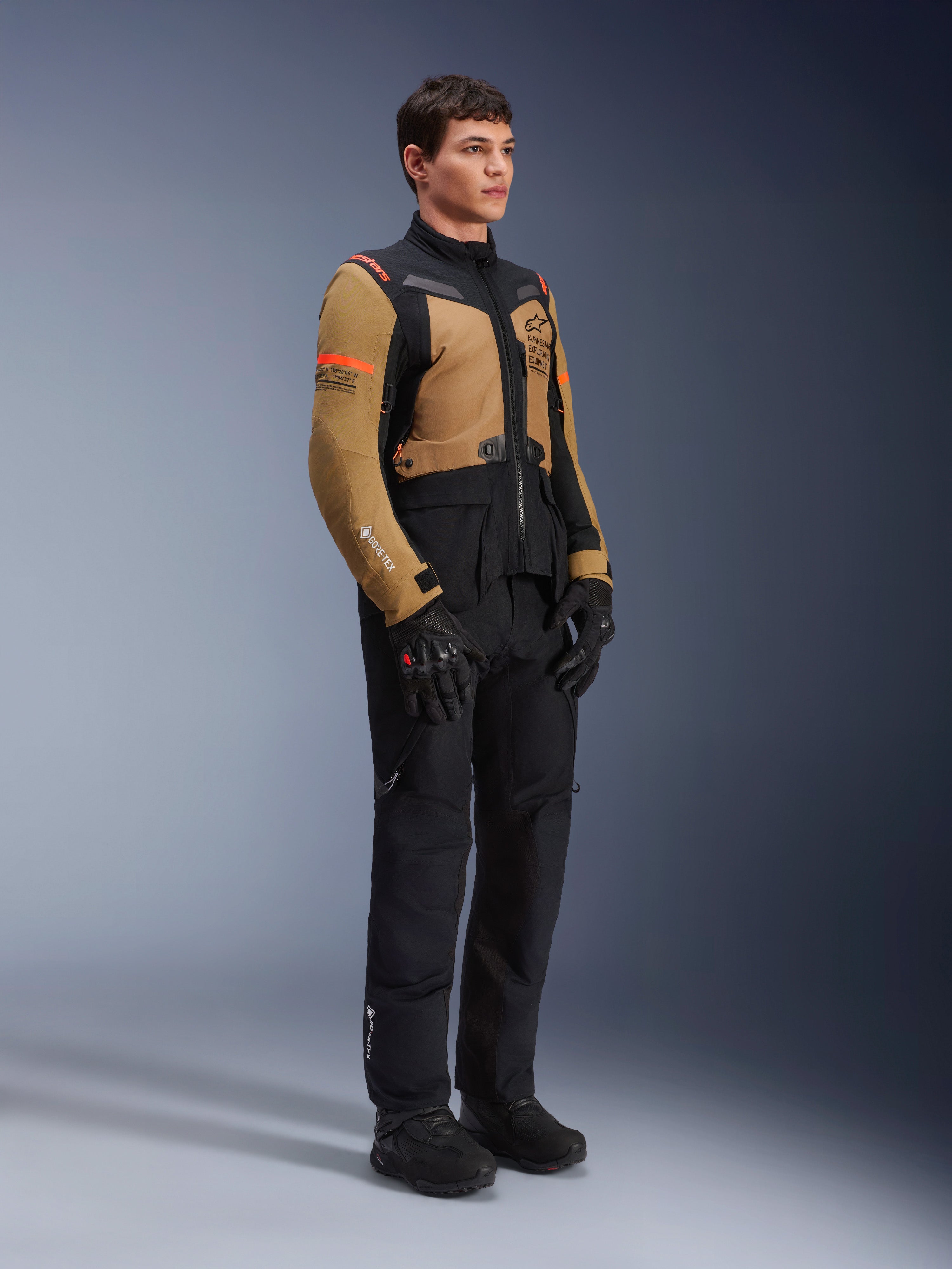 Giacca Alpinestars ST-7 2L GORE-TEX, giacca da moto, marrone Gobi e nero, indossata da un modello con pantaloni e guanti neri abbinati, con pannelli marroni sul torace e sulle maniche, accenti tecnici neri e loghi arancioni, abbigliamento da moto adventure-touring.