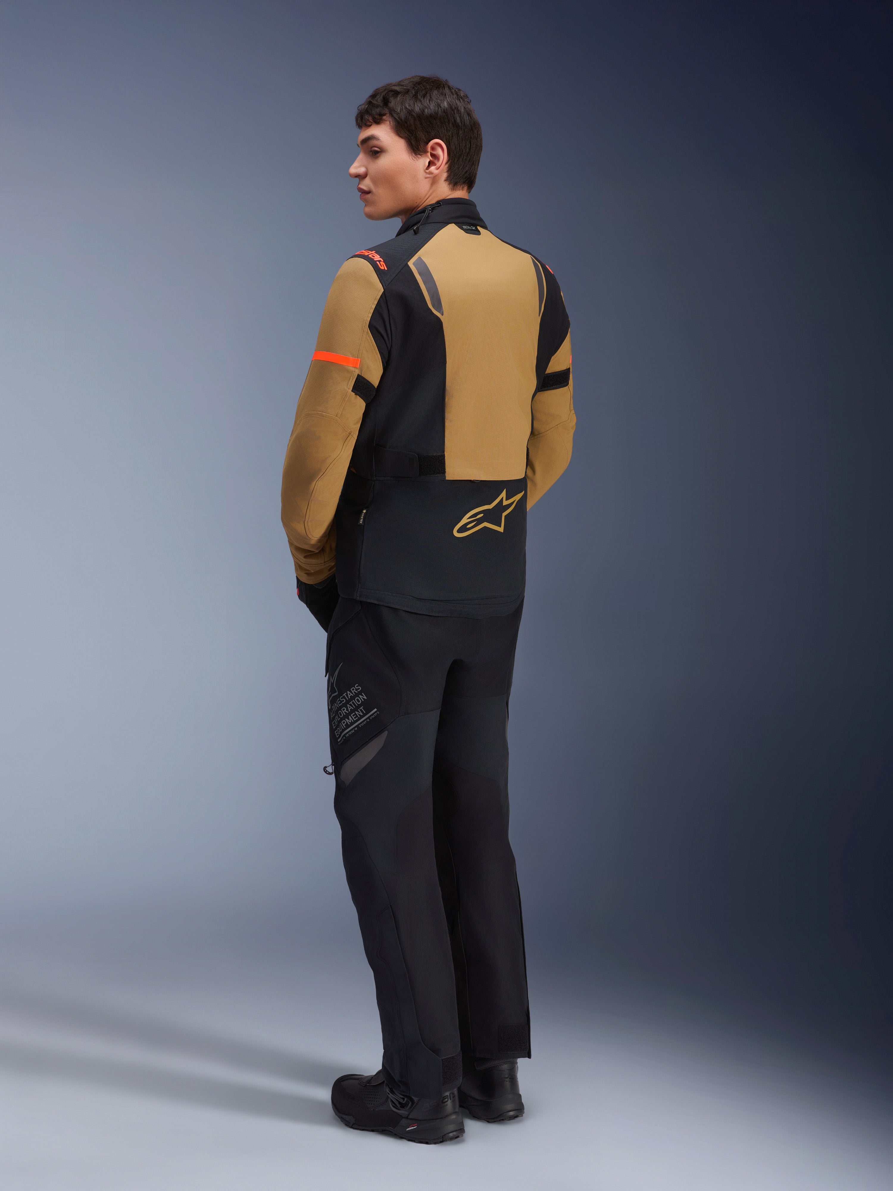 Giacca Alpinestars ST-7 2L GORE-TEX, giacca da moto adventure, Gobi Brown e nero, vista posteriore su un modello che indossa pantaloni e stivali da moto coordinati, con dettagli arancioni sulle maniche, guscio laminato impermeabile e logo Alpinestars, progettata per il touring e la guida su strada