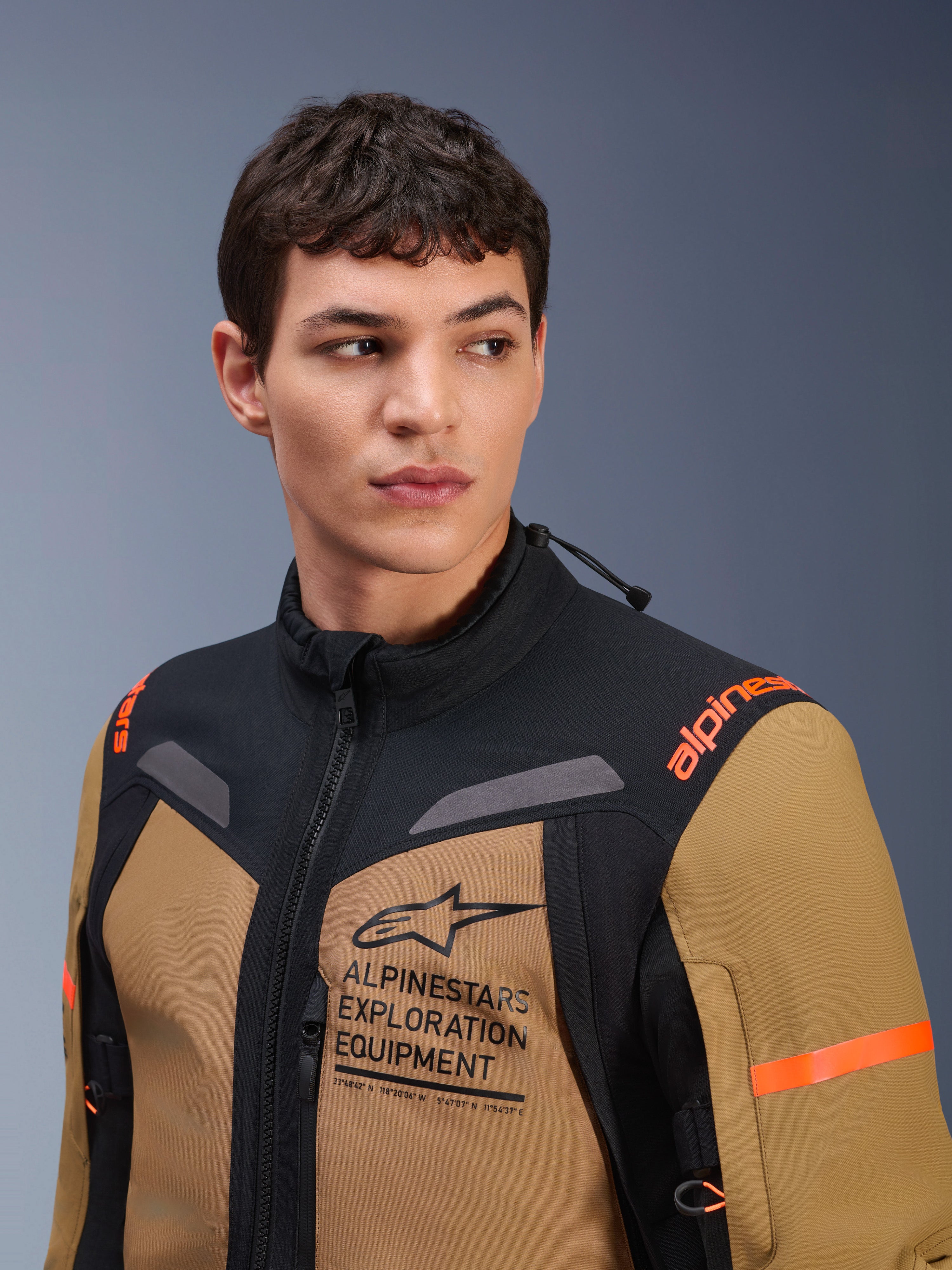 Giacca Alpinestars ST-7 2L GORE-TEX, Gobi Brown e nero, indossata da un modello uomo in uno studio, con l'evidente marchio 'ALPINESTARS EXPLORATION EQUIPMENT' sul petto, loghi arancioni sulle spalle e struttura tecnica in tessuto impermeabile per la guida adventure.