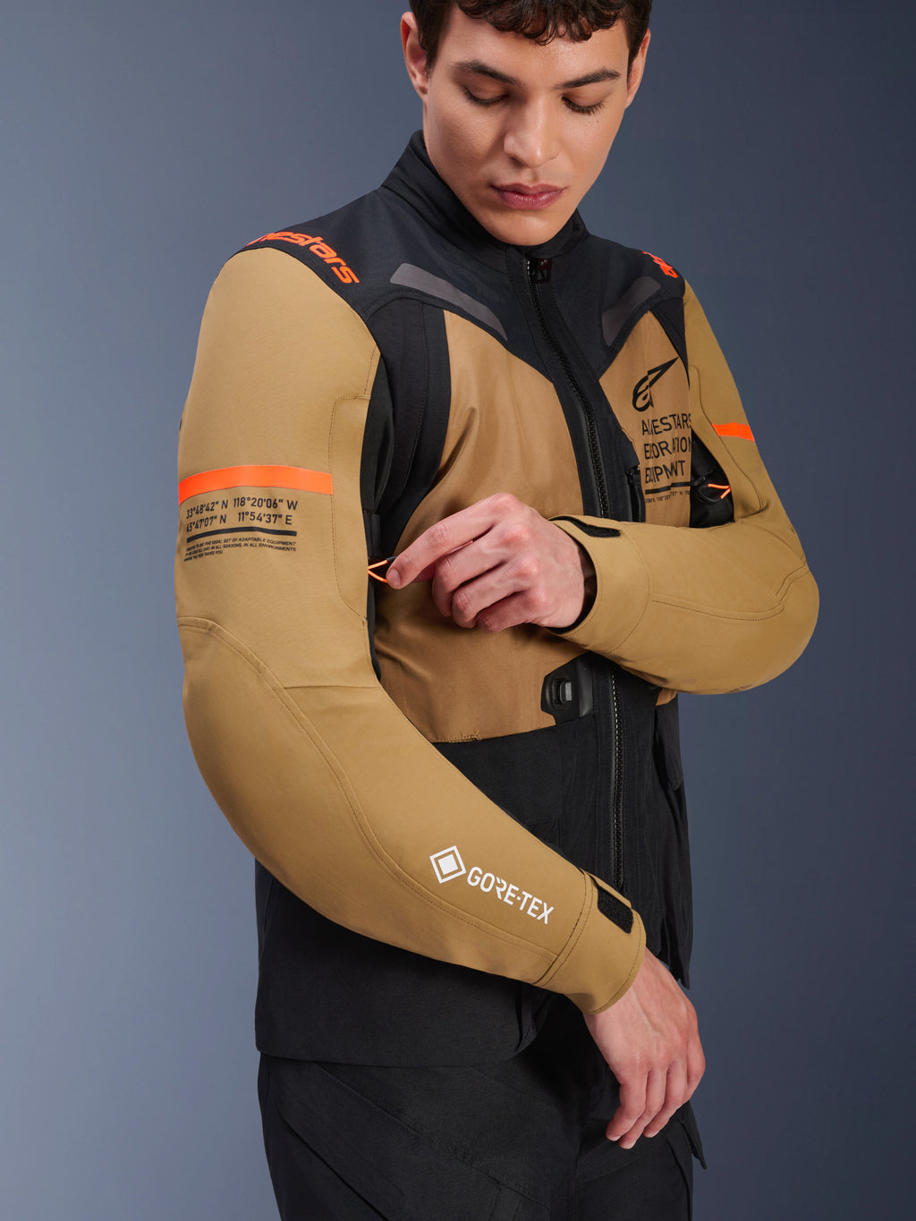 Alpinestars ST-7 2L GORE-TEX Jacket in Gobi Brown e nero, giacca da moto adventure-touring indossata da un modello che regola una presa d'aria sulla manica, con accenti arancioni, grafiche con coordinate e logo GORE-TEX