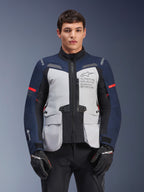 Giacca Alpinestars ST-7 2L GORE-TEX, giacca da moto adventure touring, grigio ghiaccio e blu scuro con dettagli rossi, impermeabile e traspirante, indossata da un modello con guanti neri, con logo Alpinestars Exploration Equipment sul petto
