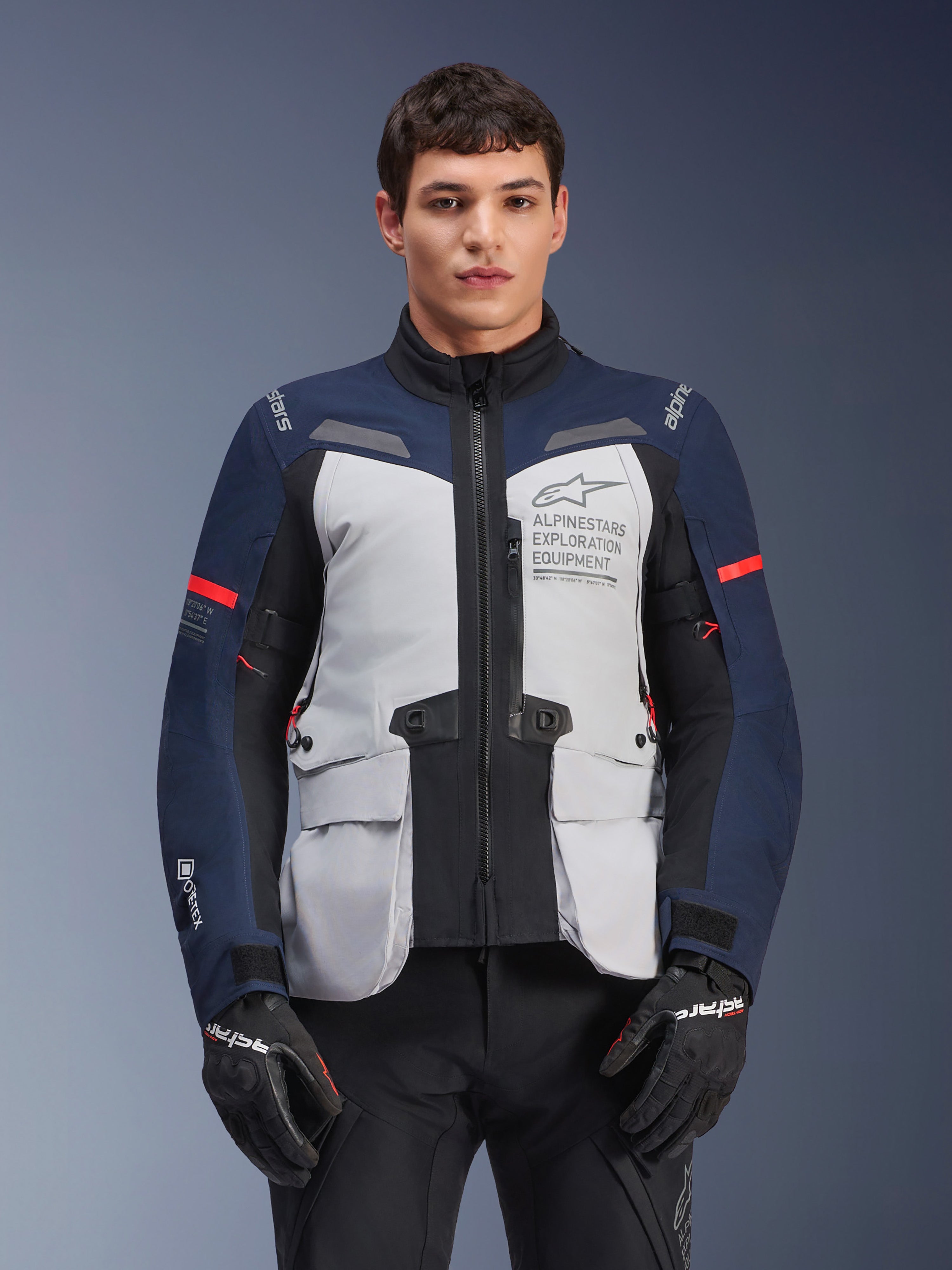 Giacca Alpinestars ST-7 2L GORE-TEX, giacca da moto adventure touring, grigio ghiaccio e blu scuro con dettagli rossi, impermeabile e traspirante, indossata da un modello con guanti neri, con logo Alpinestars Exploration Equipment sul petto