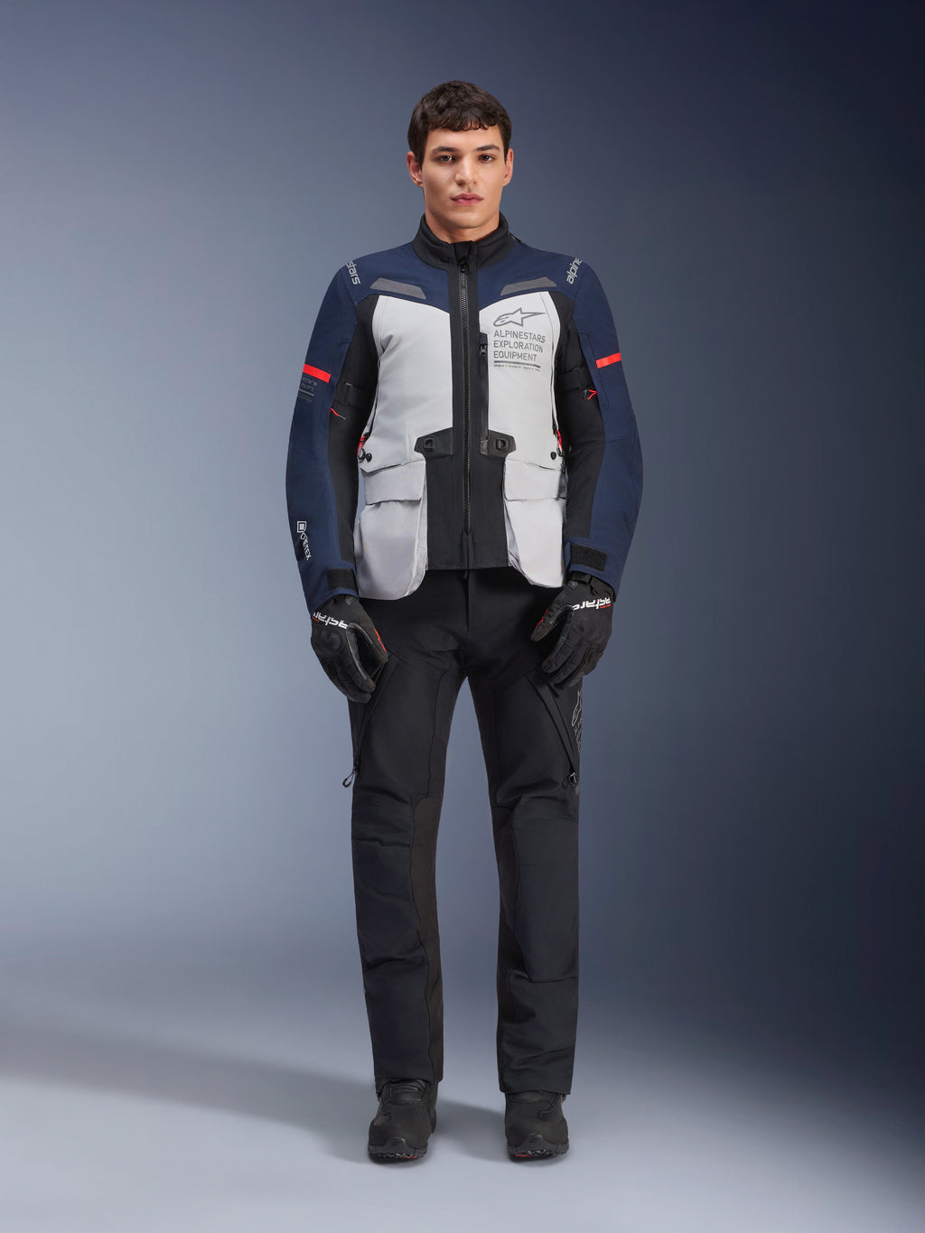 Giacca Alpinestars ST-7 2L GORE-TEX, giacca da moto Adventure Touring, Grigio Ghiaccio, Blu Scuro e Nero, indossata con pantaloni e guanti neri, dotata di ampie tasche cargo anteriori, dettagli rossi sulle maniche e guscio in GORE-TEX laminato per prestazioni impermeabili.