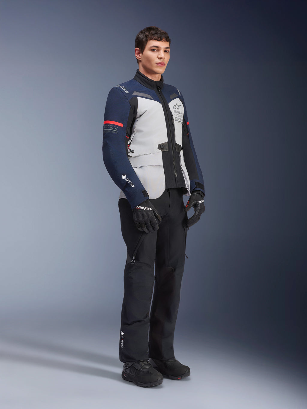 Giacca Alpinestars ST-7 2L GORE-TEX, colorazione Ice Gray, blu scuro e nero con accenti rossi, indossata da un modello con pantaloni da moto e guanti neri, abbigliamento professionale da moto adventure touring