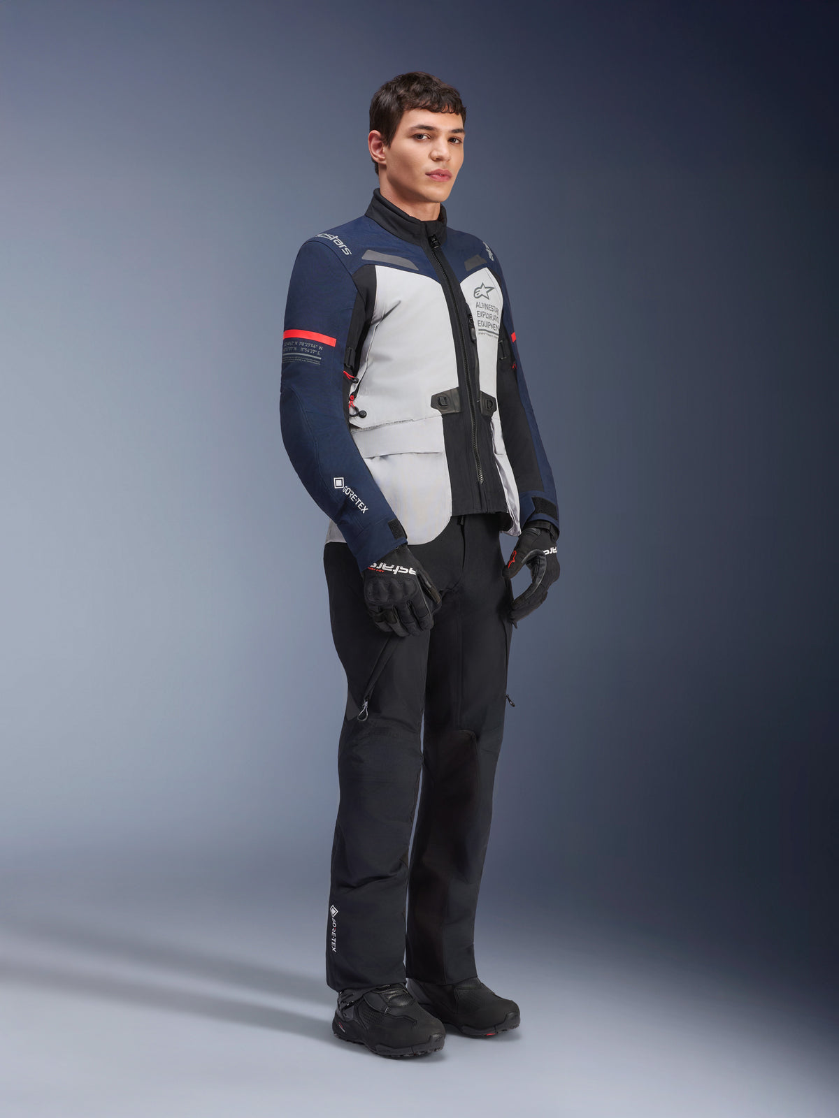 Giacca Alpinestars ST-7 2L GORE-TEX, colorazione Ice Gray, blu scuro e nero con accenti rossi, indossata da un modello con pantaloni da moto e guanti neri, abbigliamento professionale da moto adventure touring