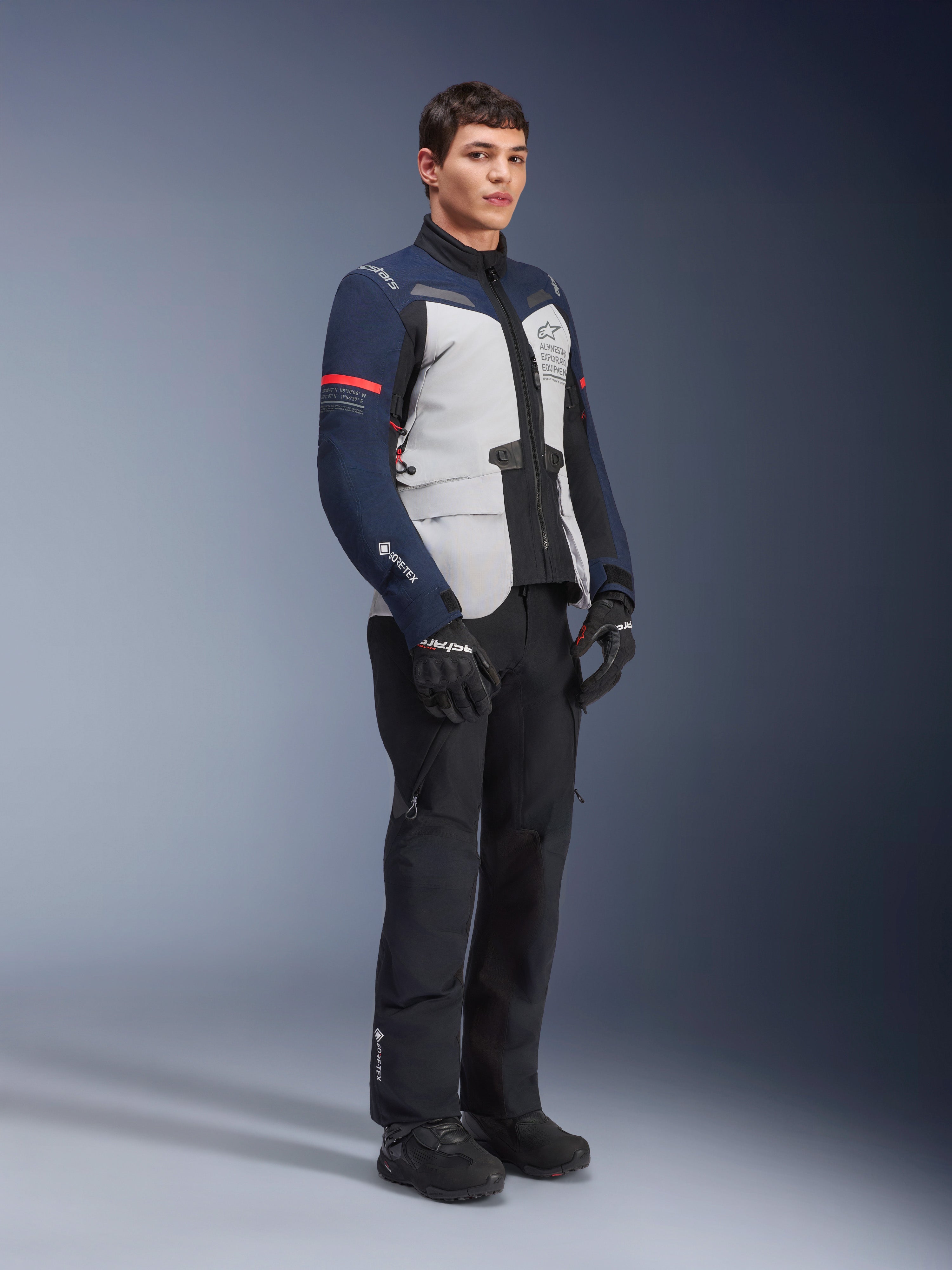 Giacca Alpinestars ST-7 2L GORE-TEX, colorazione Ice Gray, blu scuro e nero con accenti rossi, indossata da un modello con pantaloni da moto e guanti neri, abbigliamento professionale da moto adventure touring