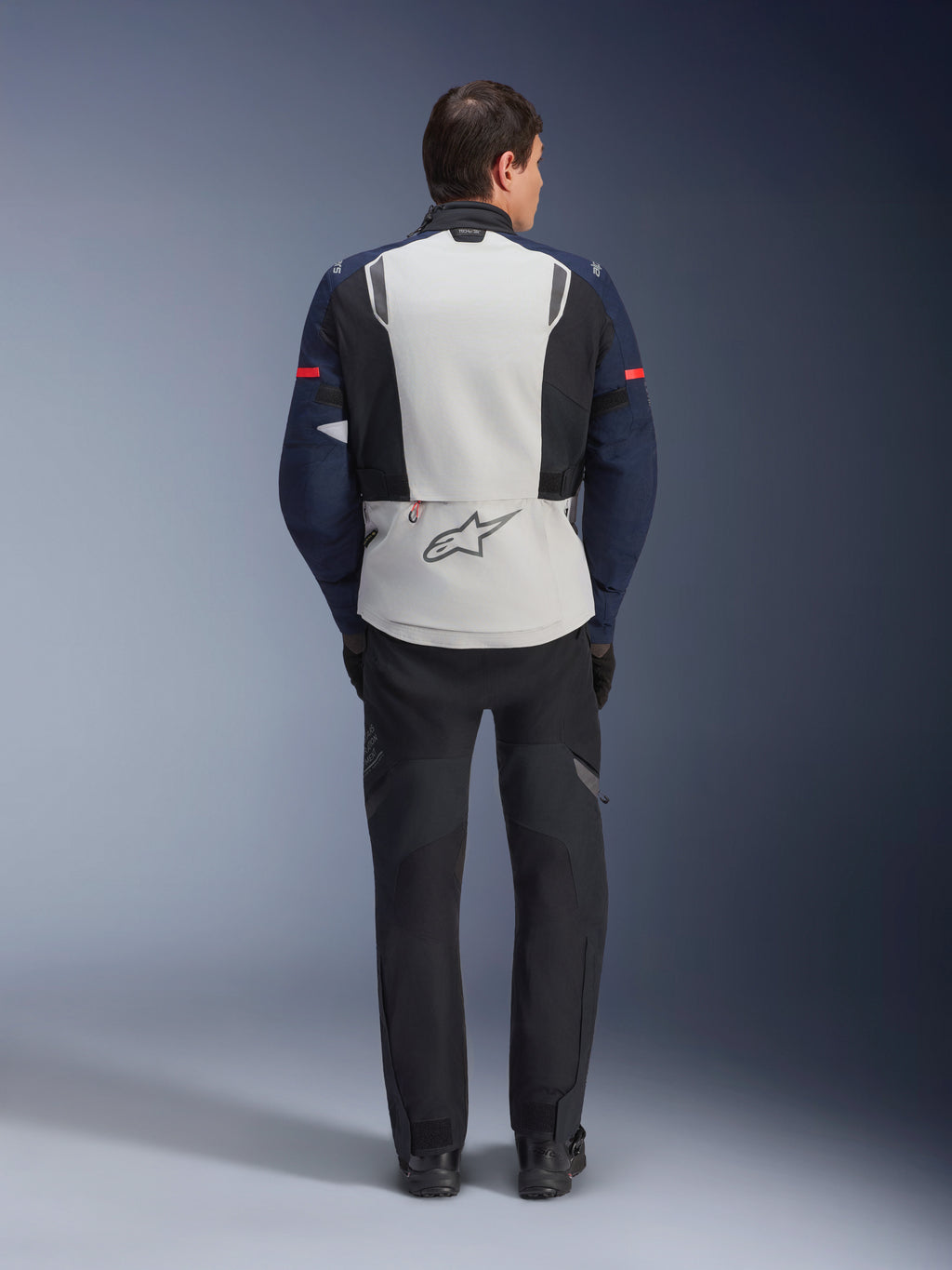 Giacca Alpinestars ST-7 2L GORE-TEX, vista posteriore di un pilota che indossa una giacca da moto adventure touring in Ice Gray, Dark Blue e nero, con ampio pannello posteriore, logo Alpinestars e pantaloni neri abbinati, progettata per ogni condizione meteorologica.